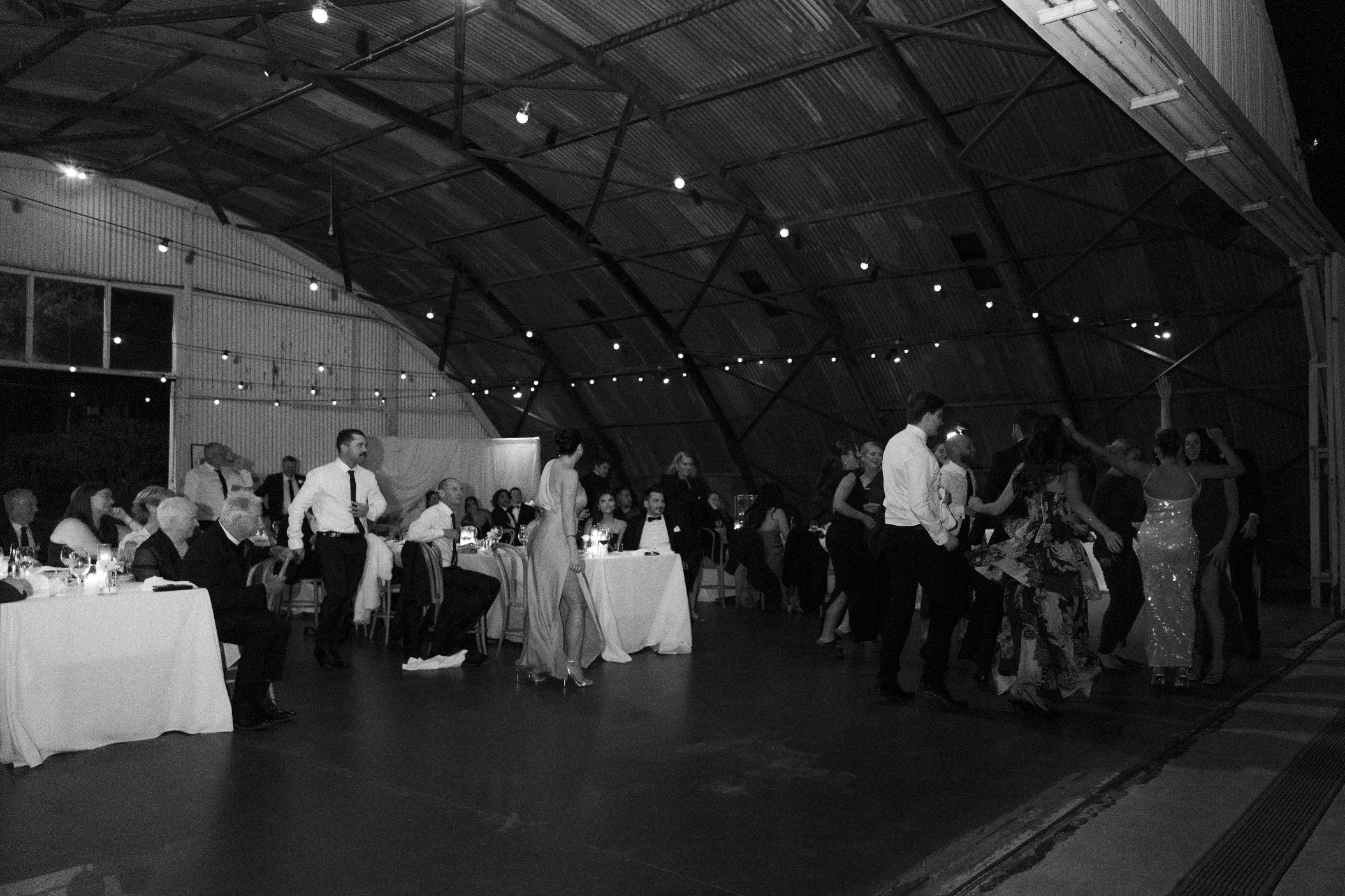 People dancing and socializing in a large indoor event space with a high, curved ceiling and hanging string lights, at what appears to be a wedding reception or celebration.