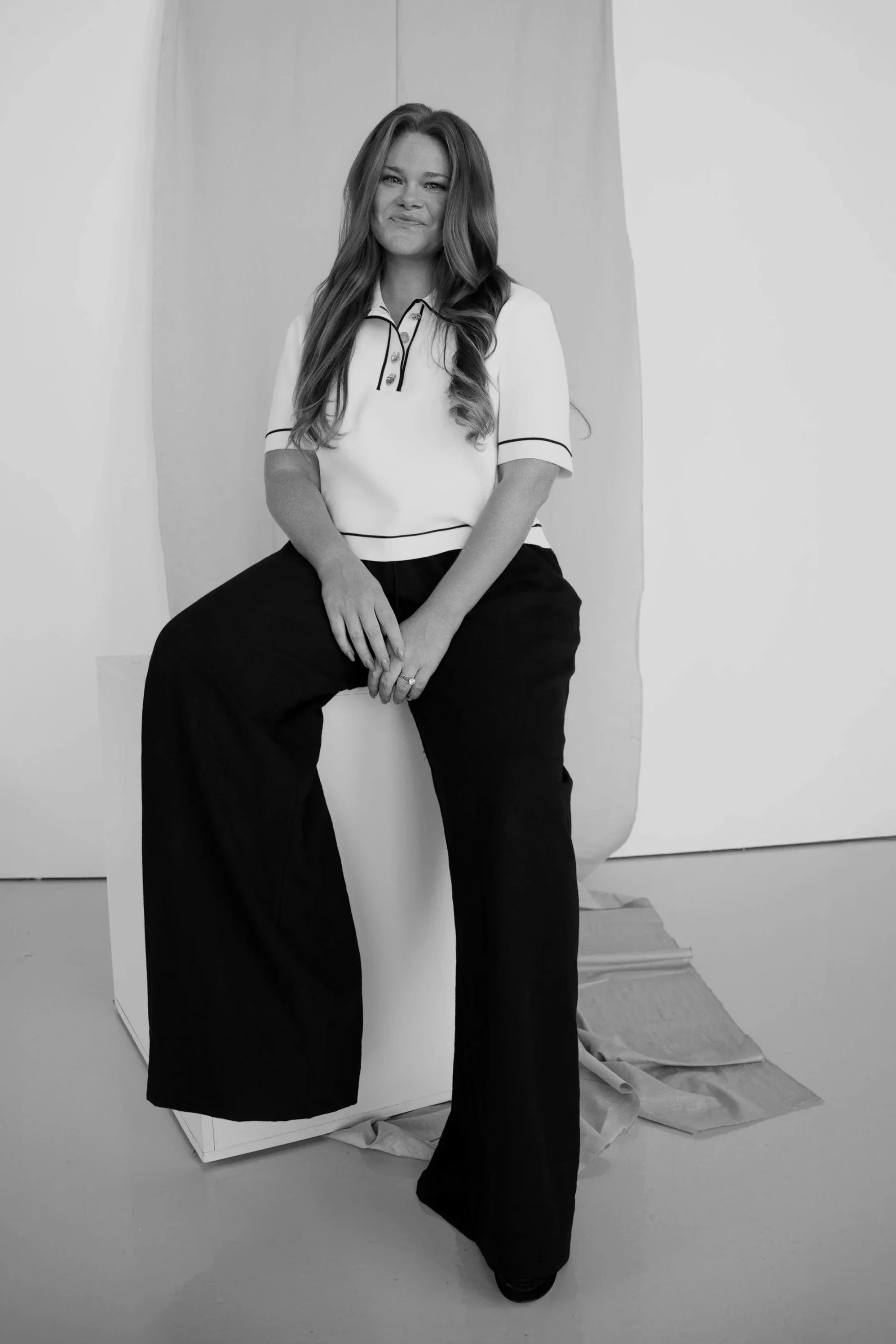Black and white photo of a woman sitting in front of a plain background. She has long wavy hair and is wearing a short-sleeved shirt with dark pants. She is smiling and has her hands resting on her lap.