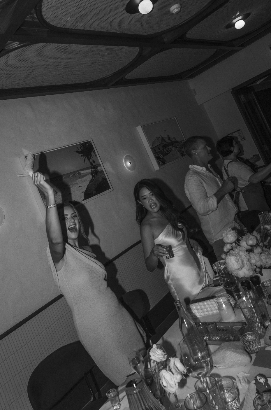 People celebrating at a party with drinks and flowers on the table, women dancing and raising their hands, in a room with pictures on the wall.