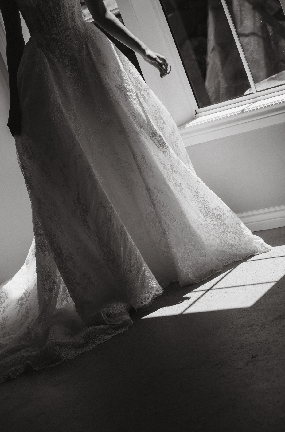 A bride in a lace wedding gown standing by a window, with sunlight casting shadows on the floor.