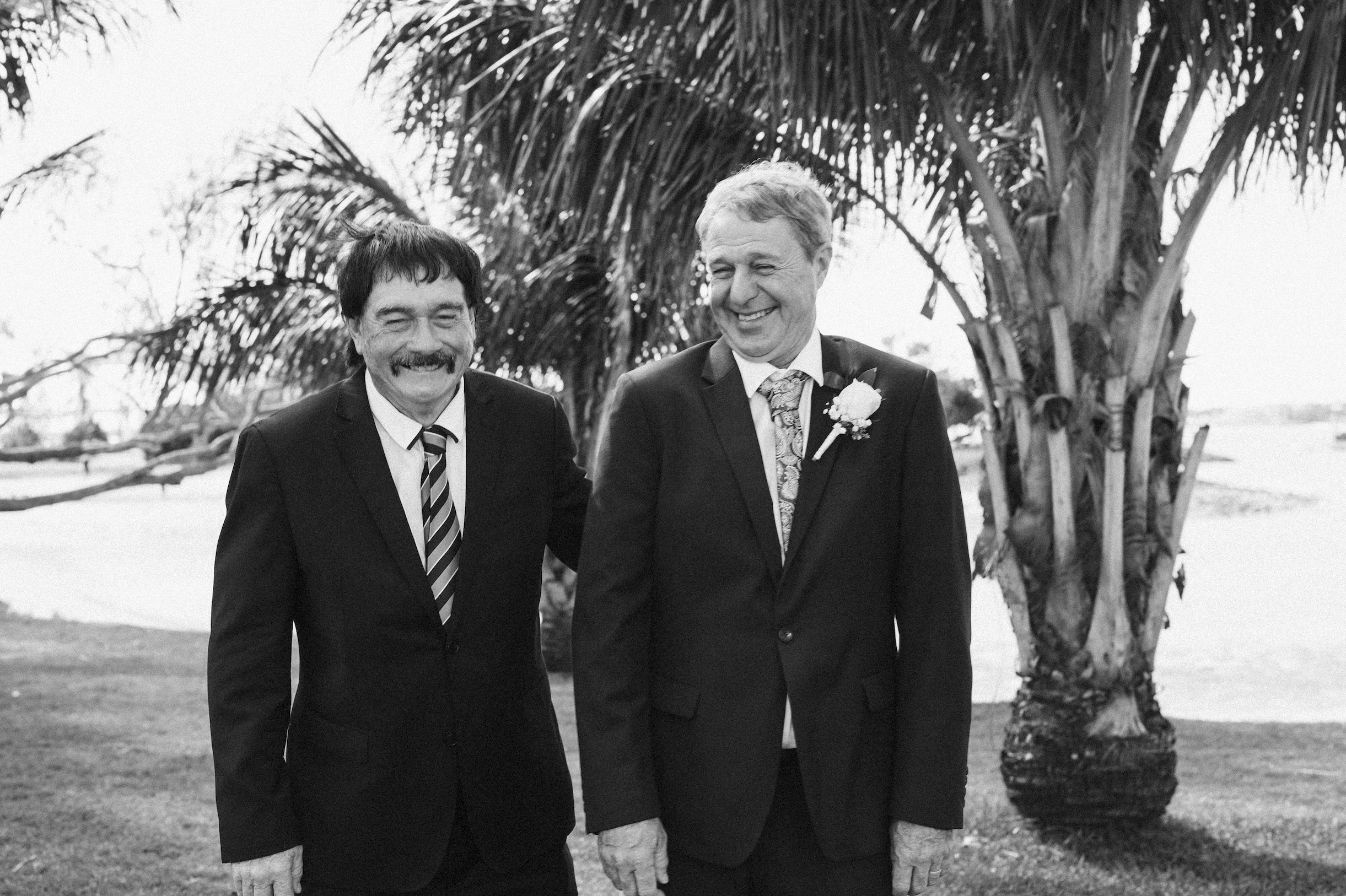 Two men dressed in suits standing outdoors near a palm tree, smiling and laughing, in black and white photograph.