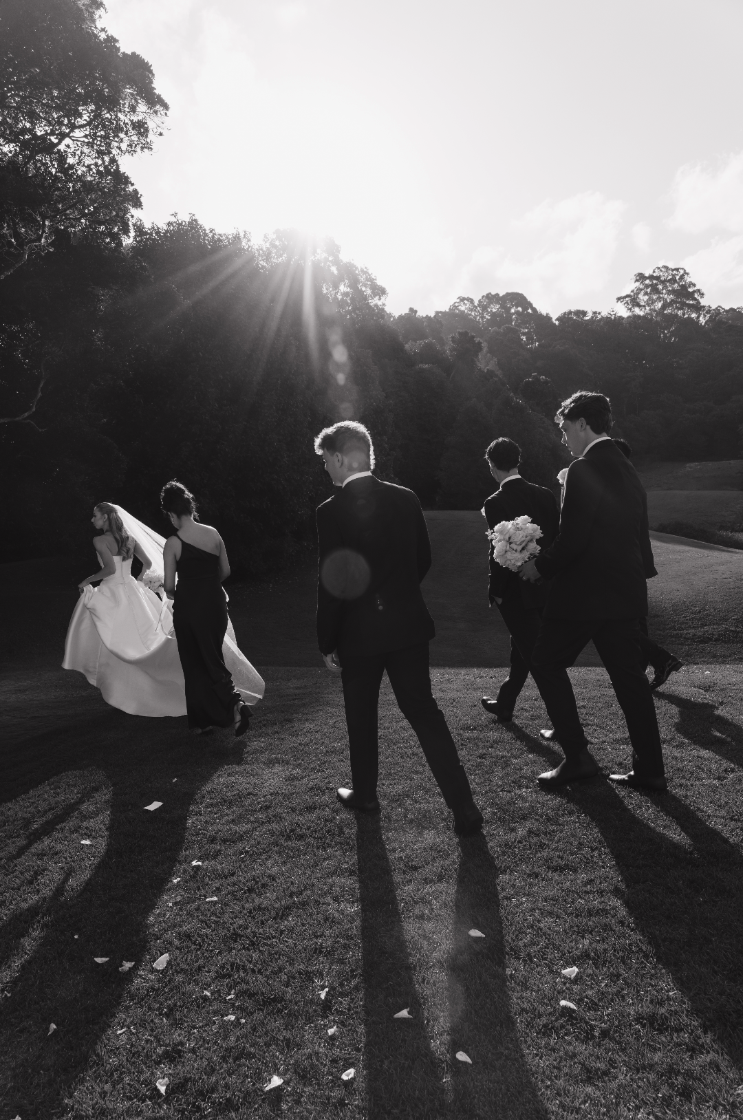 Black and white photo of a wedding party walking outdoors on a grassy area, with the bride in a white dress and veil, and five men in suits, one holding a bouquet of flowers, as the sun shines overhead, casting long shadows.
