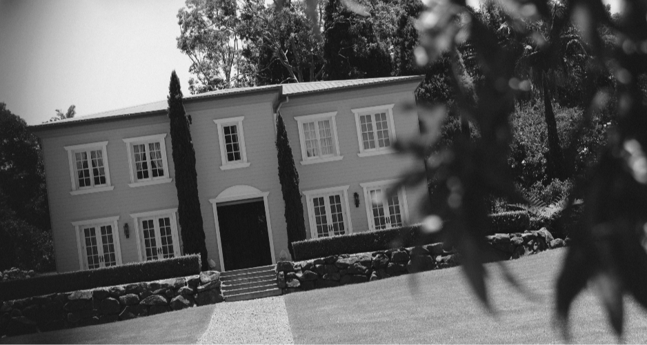 A two-story house with a symmetrical façade, surrounded by trees. The house has a central entrance with stairs, tall narrow windows, and appears to be styled in traditional architecture. Black and white image.