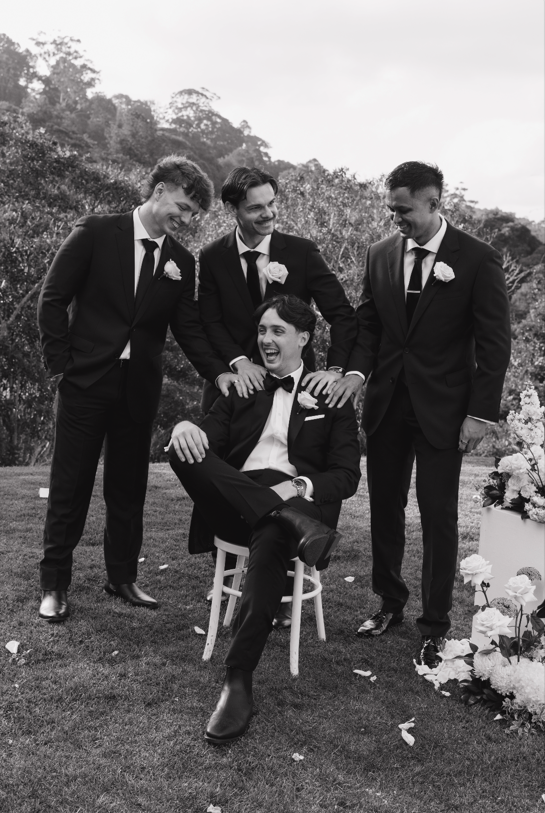 Four men dressed in tuxedos, with one sitting on a chair and three standing around him outdoors, smiling and laughing during a social gathering or wedding celebration in a natural setting with trees in the background.
