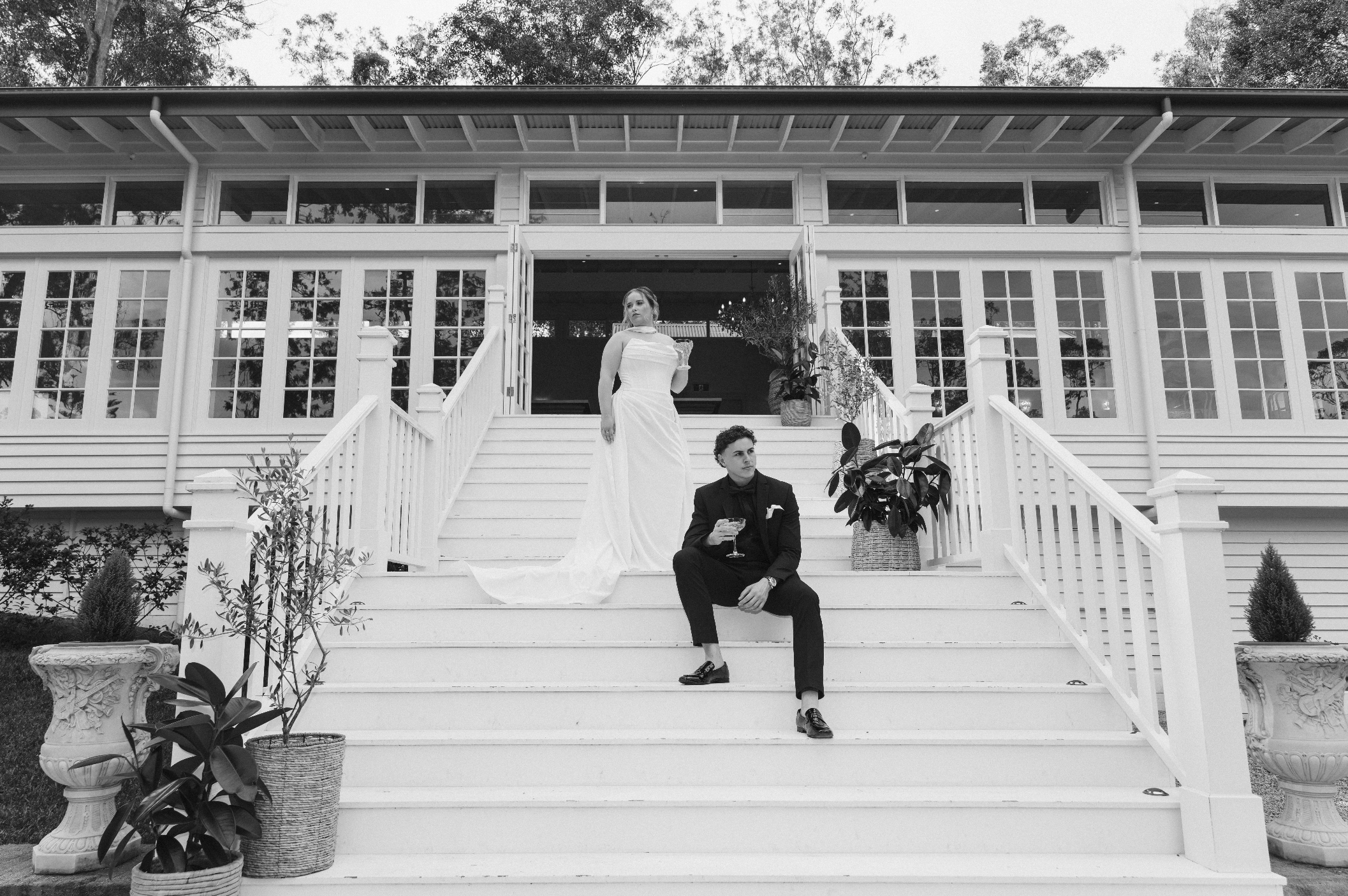 A black and white photo of a woman in a white wedding dress and a man in a tuxedo sitting on the stairs of a large white house with glass railings, plants, and trees in the background.