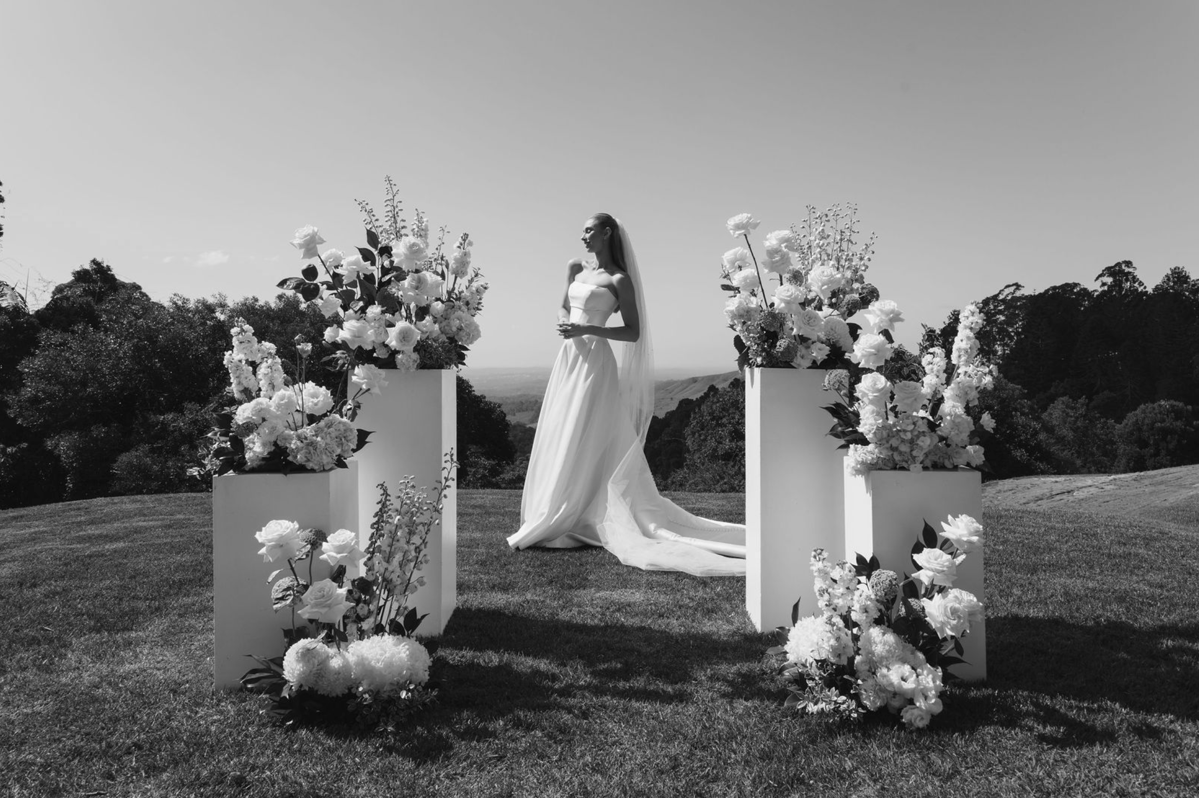 A black and white photo of a woman in a wedding dress standing outdoors with mountains and trees in the background, surrounded by large floral arrangements on white pedestals.