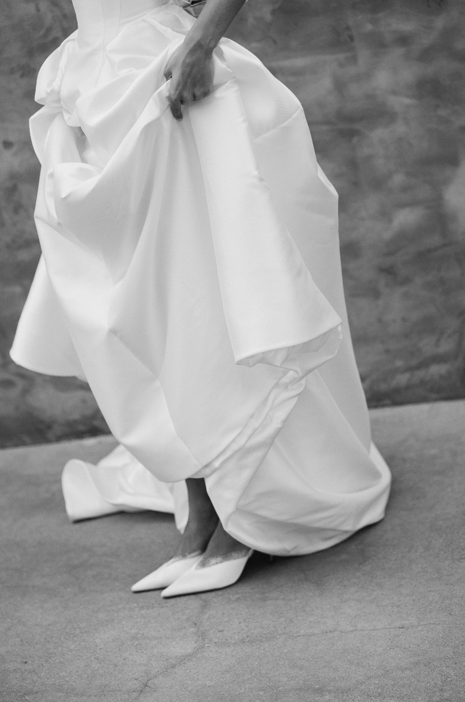 Close-up of a person wearing a flowing white dress and white high heels, holding the dress with one hand, standing on a plain floor with a textured wall background.