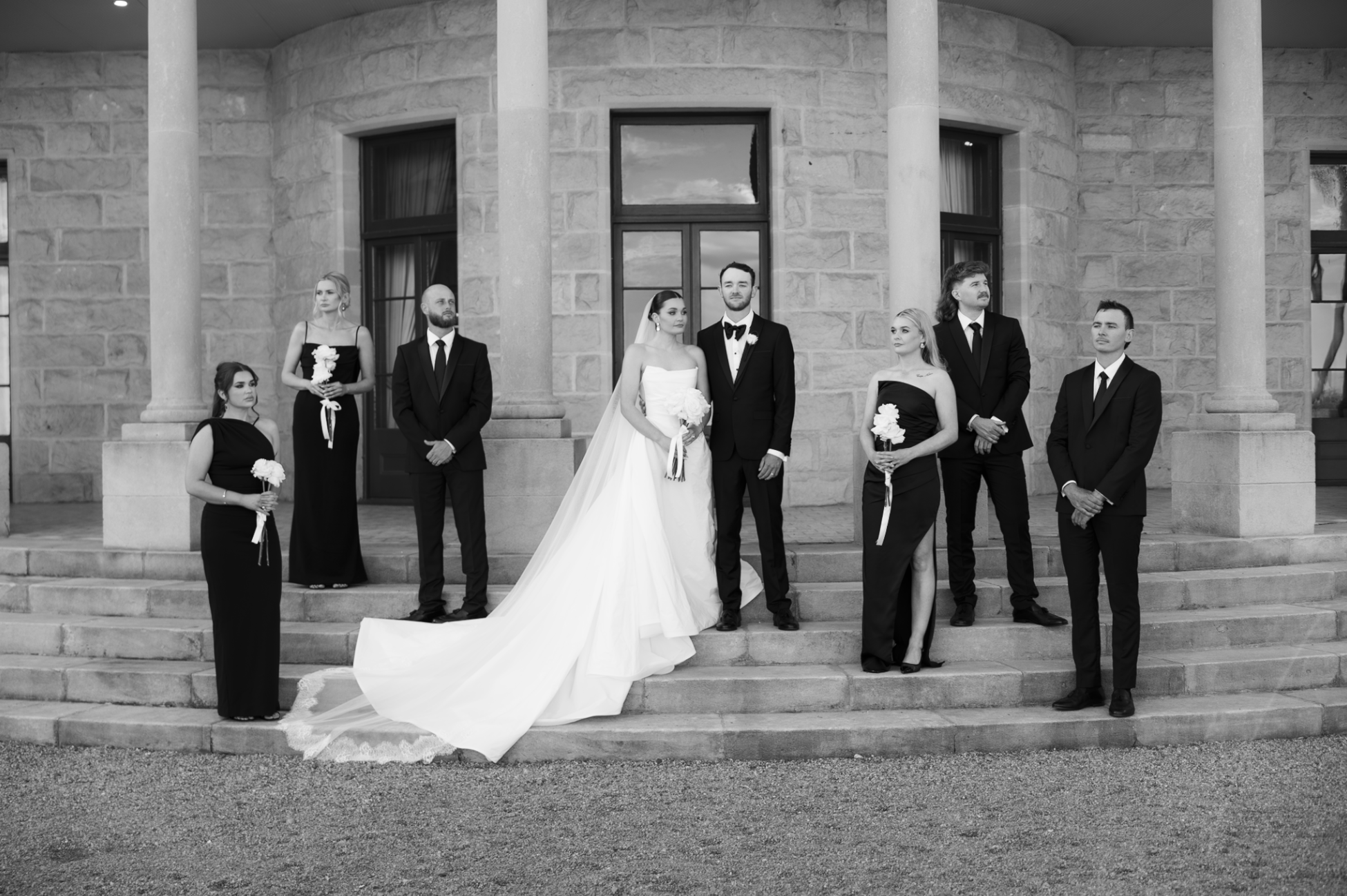 A wedding party standing on the steps of a stone building with large columns. The bride and groom are in the center, with the bride wearing a white wedding dress and the groom in a black tuxedo. The bridesmaids are dressed in black gowns holding white bouquets, and the groomsmen are in black suits.