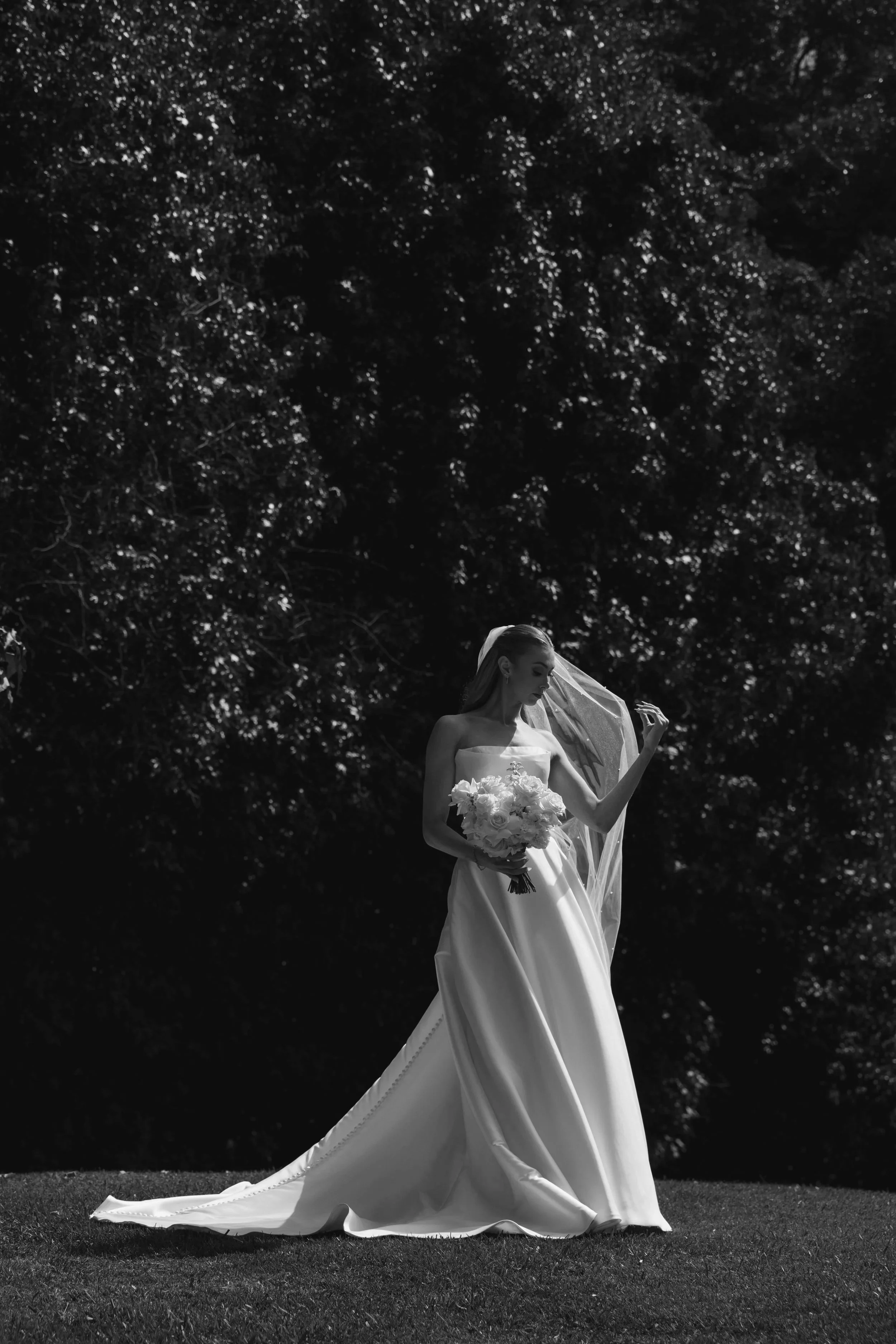 A bride in a white wedding dress holding a bouquet of flowers stands outdoors beneath tall trees, with a veil flowing behind her, captured in a black and white photograph.
