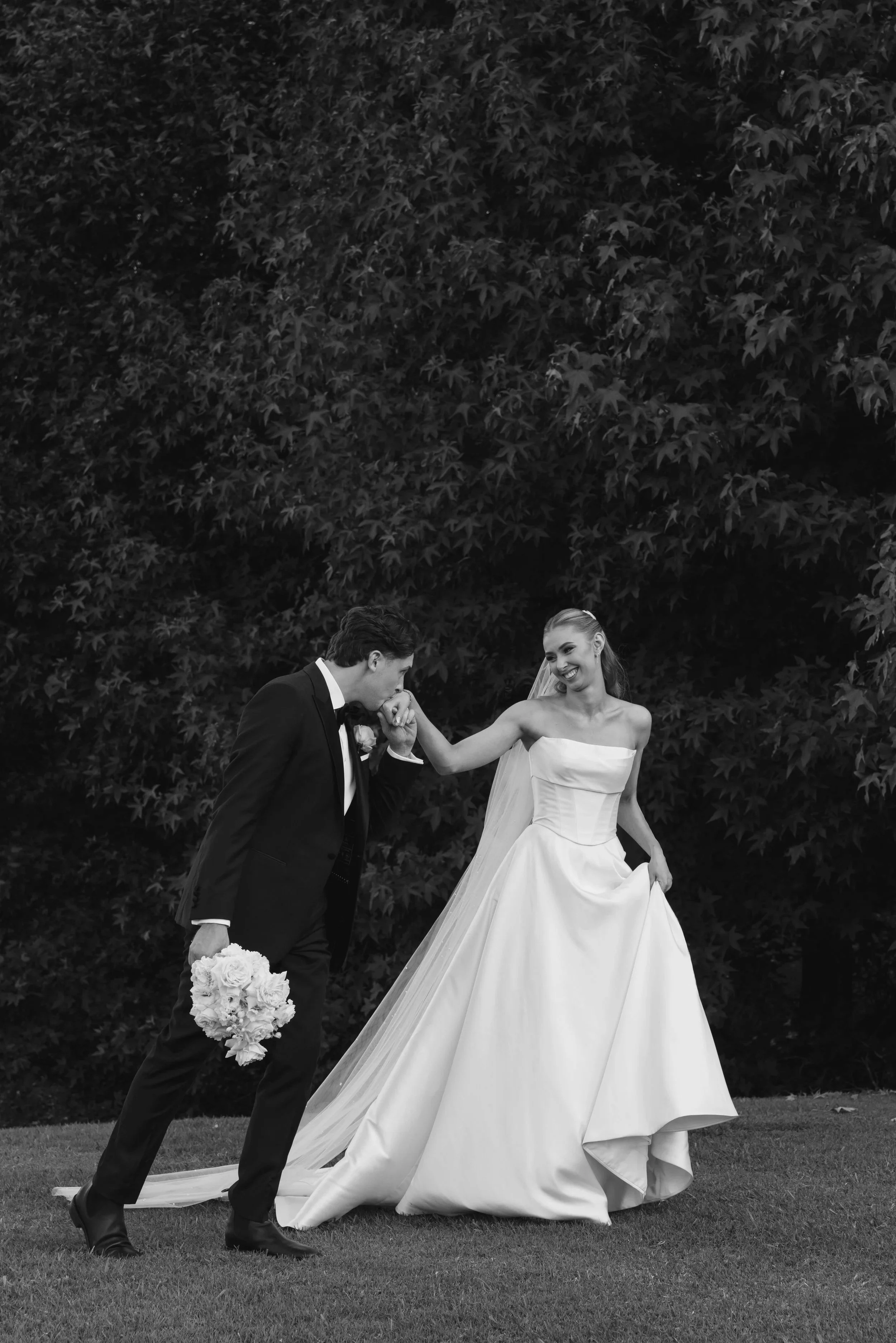 A black and white photo of a bride and groom outside, with the groom holding a bouquet of flowers and the bride smiling, wearing a wedding gown, in front of large leafy trees.