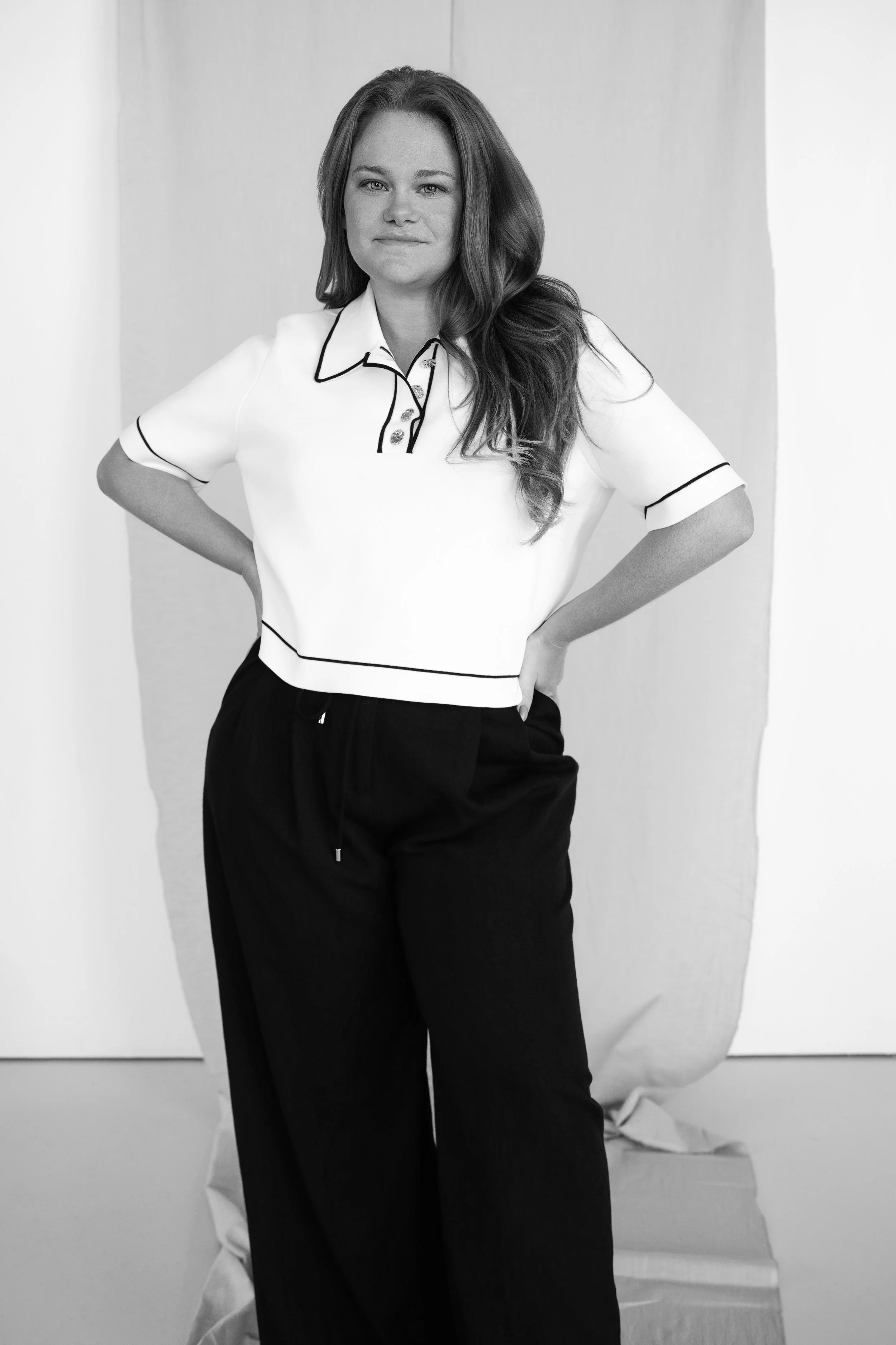 Black and white photo of a woman with long hair wearing a polo shirt and loose pants, standing with her hands on her hips in front of a backdrop with a paper or fabric draped behind her.