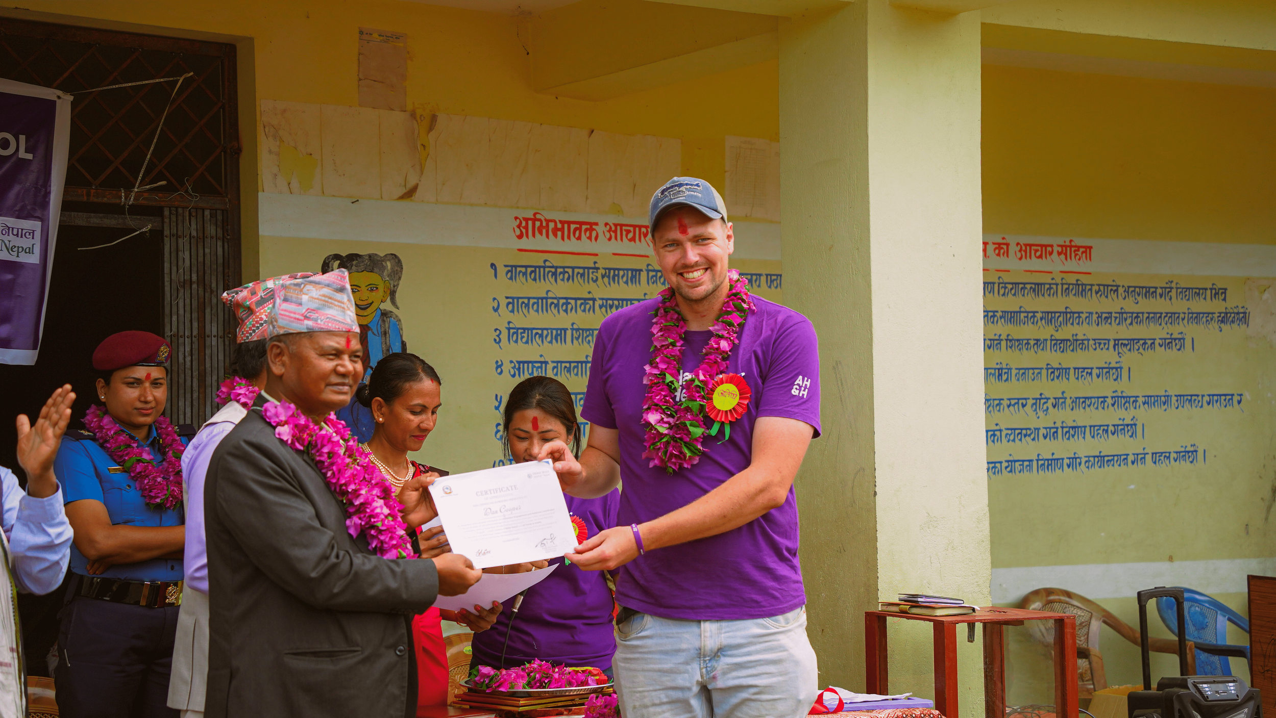 A man receiving a certificate from a woman during a ceremony, with a group of people wearing traditional Nepali attire and flower garlands, in front of a wall with text in Nepali.
