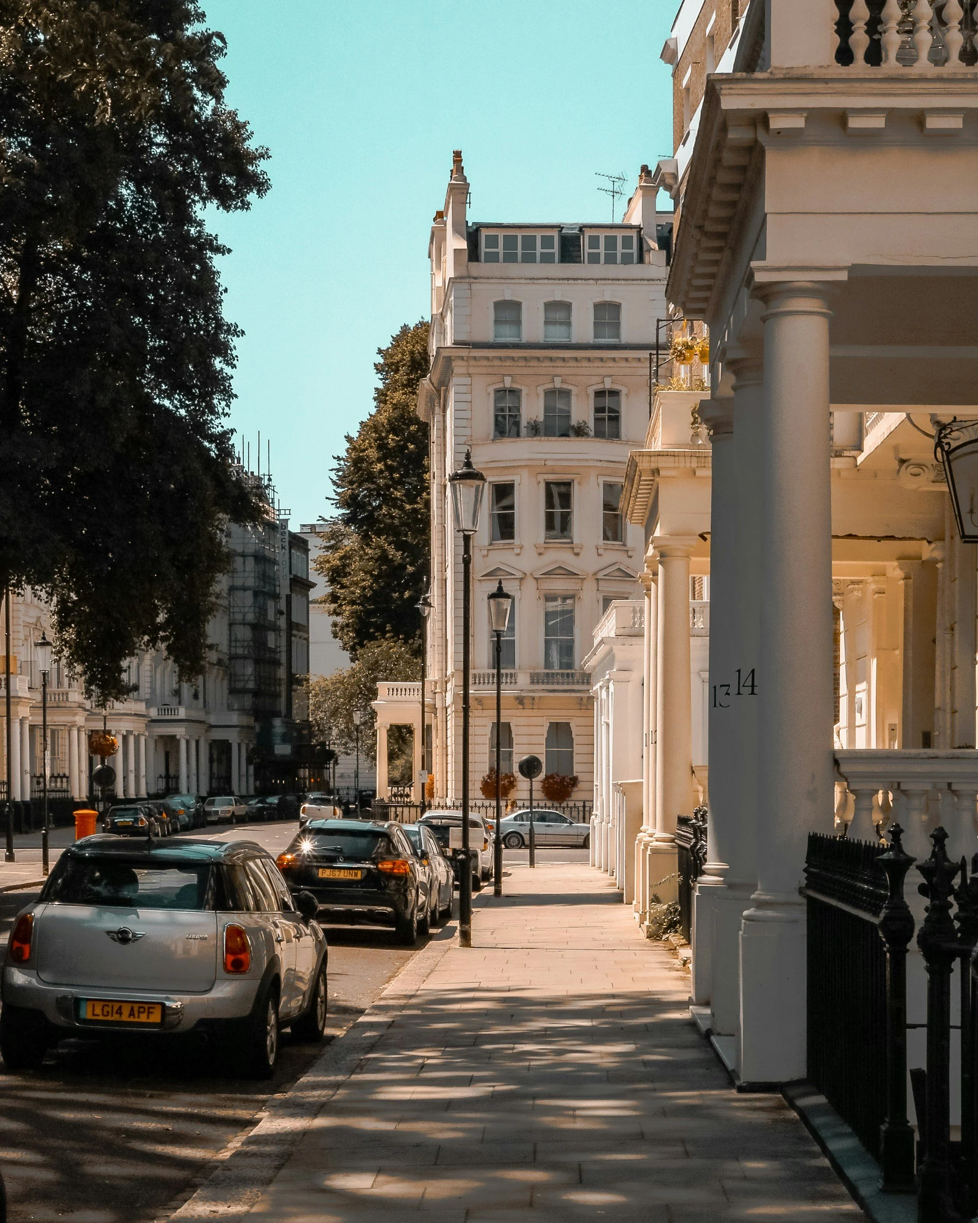 Street view in a residential area with white row houses, cars parked along the street, and a sidewalk shaded by trees in the late afternoon.