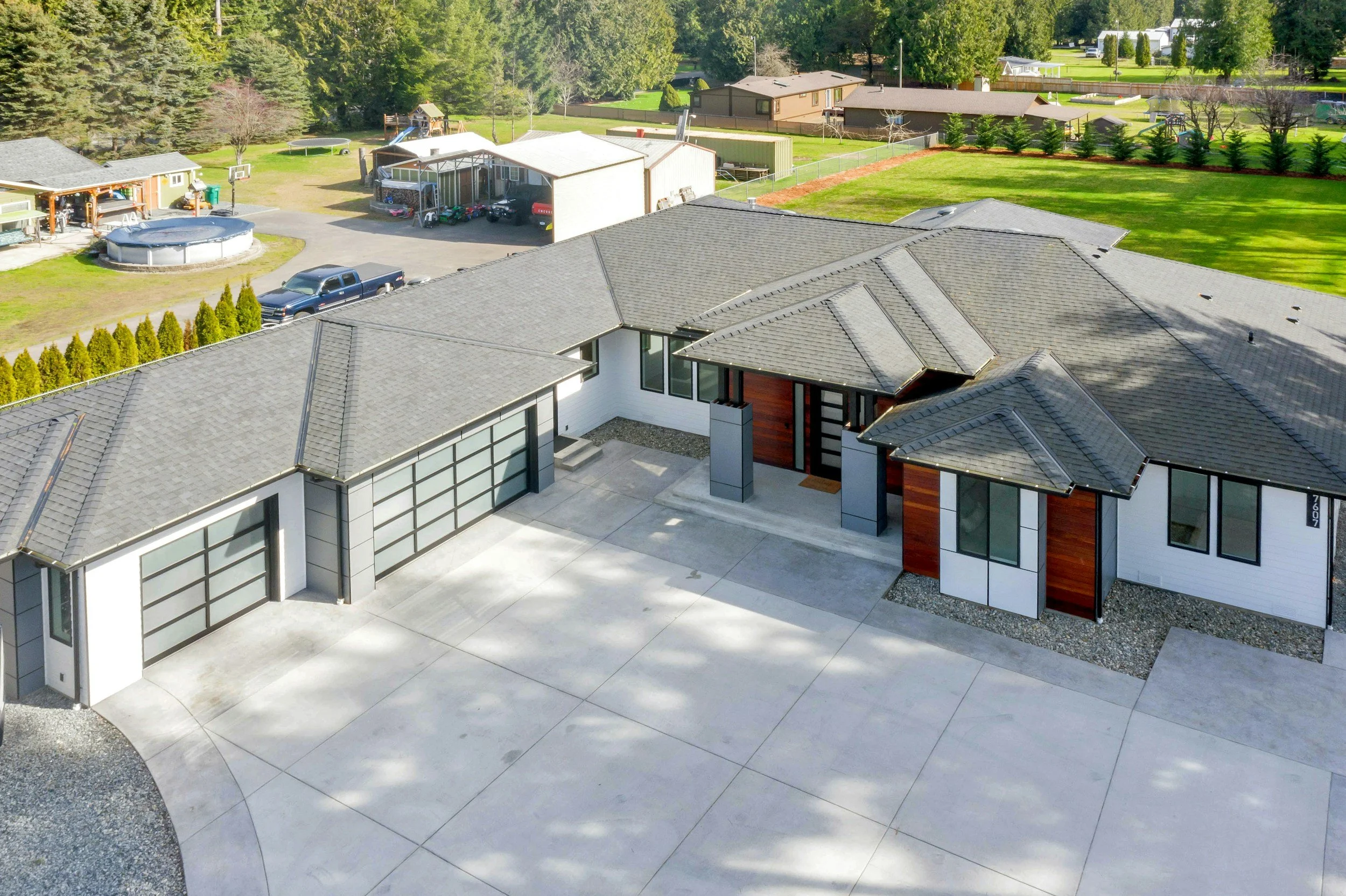 Modern house with a concrete driveway, large windows, and a landscaped yard with green grass and trees. The background shows neighboring houses, trees, and a fenced backyard.