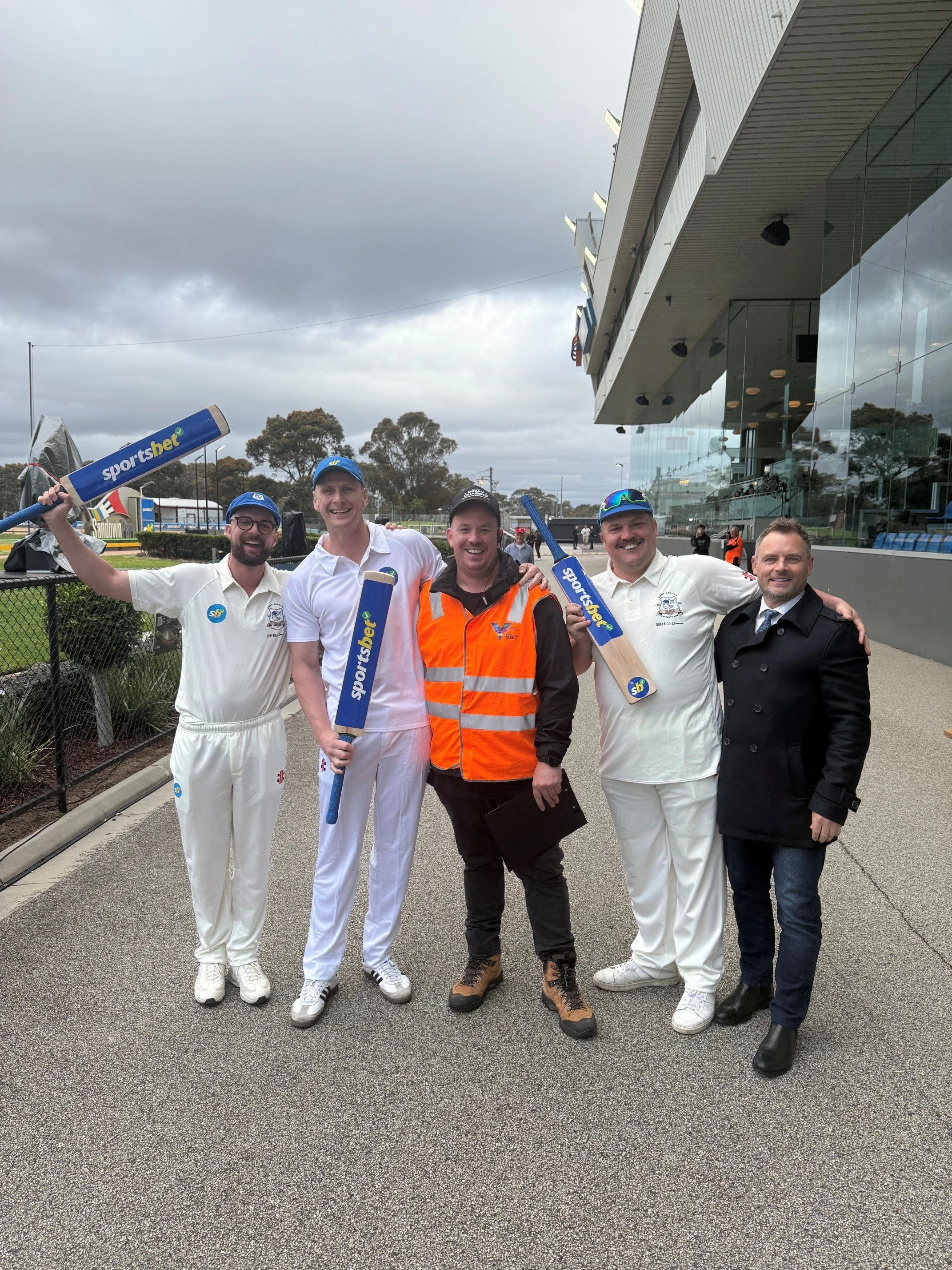 A group of five men standing outdoors, smiling, with three of them holding cricket bats. The men are wearing white cricket uniforms, one man is in an orange reflective vest, and another is in a black jacket. They are standing on a paved area near a sports building with glass windows, and there are trees and cloudy skies in the background.