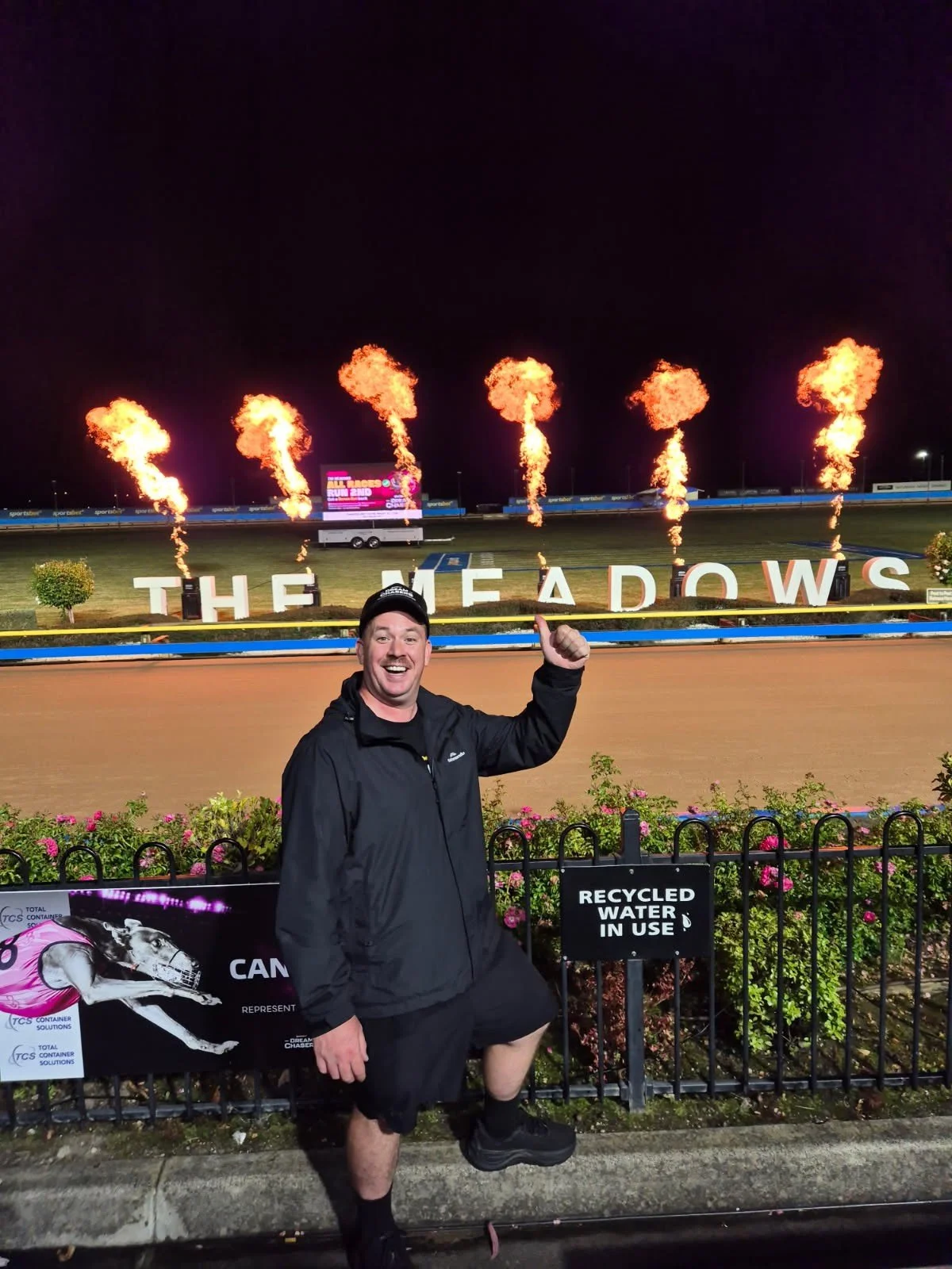 A man in black workout attire smiling and giving a thumbs-up outdoors at night, in front of a sign that reads 'THE MEADOWS' with flames shooting into the sky behind it.
