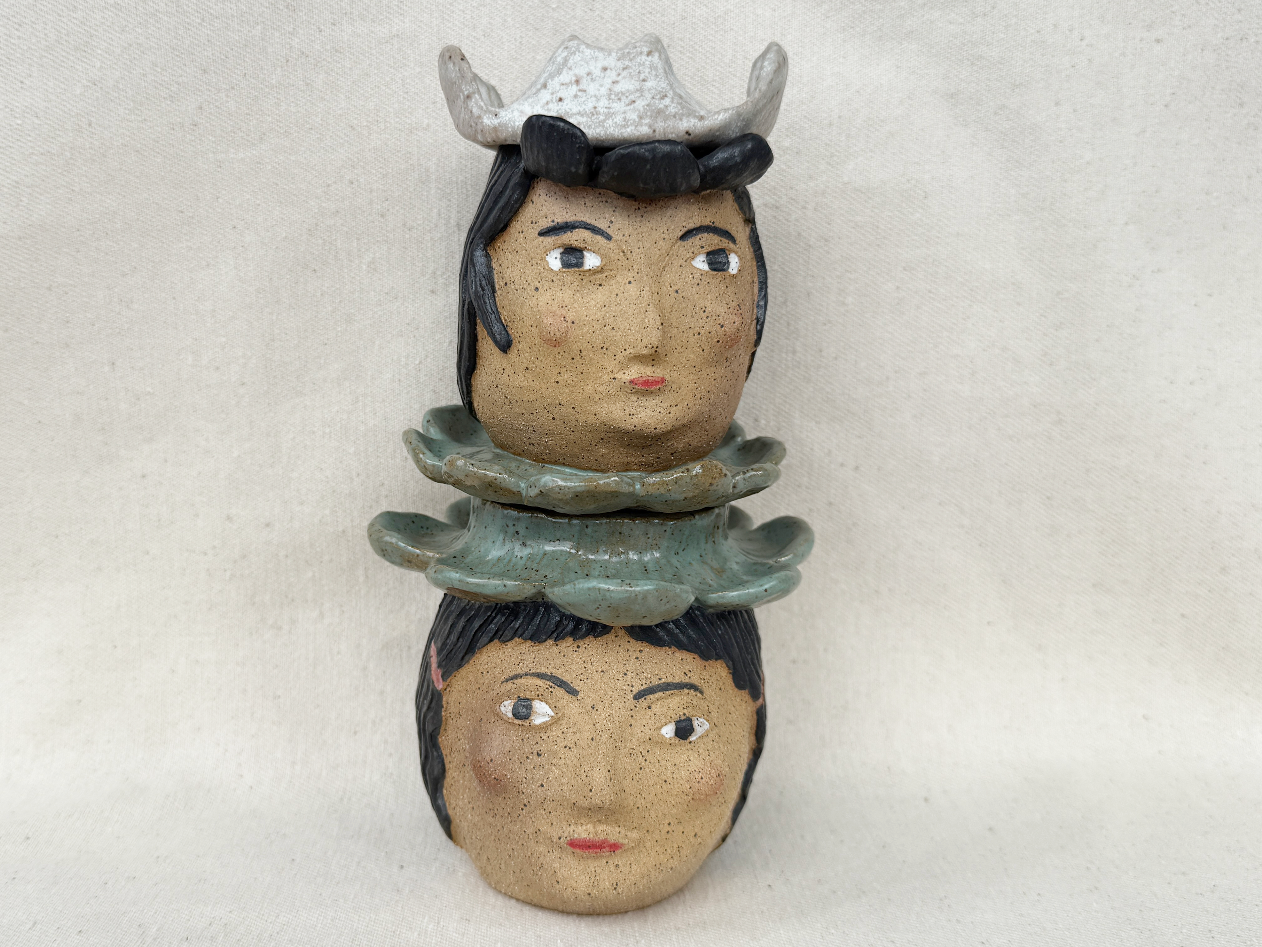 A ceramic sculpture of a woman's face with black hair, painted eyes, and red lips, wearing a large, ornate collar. The statue has a textured surface and is topped with a crown made of rocks and a white shell. It is set against a plain light backgroun