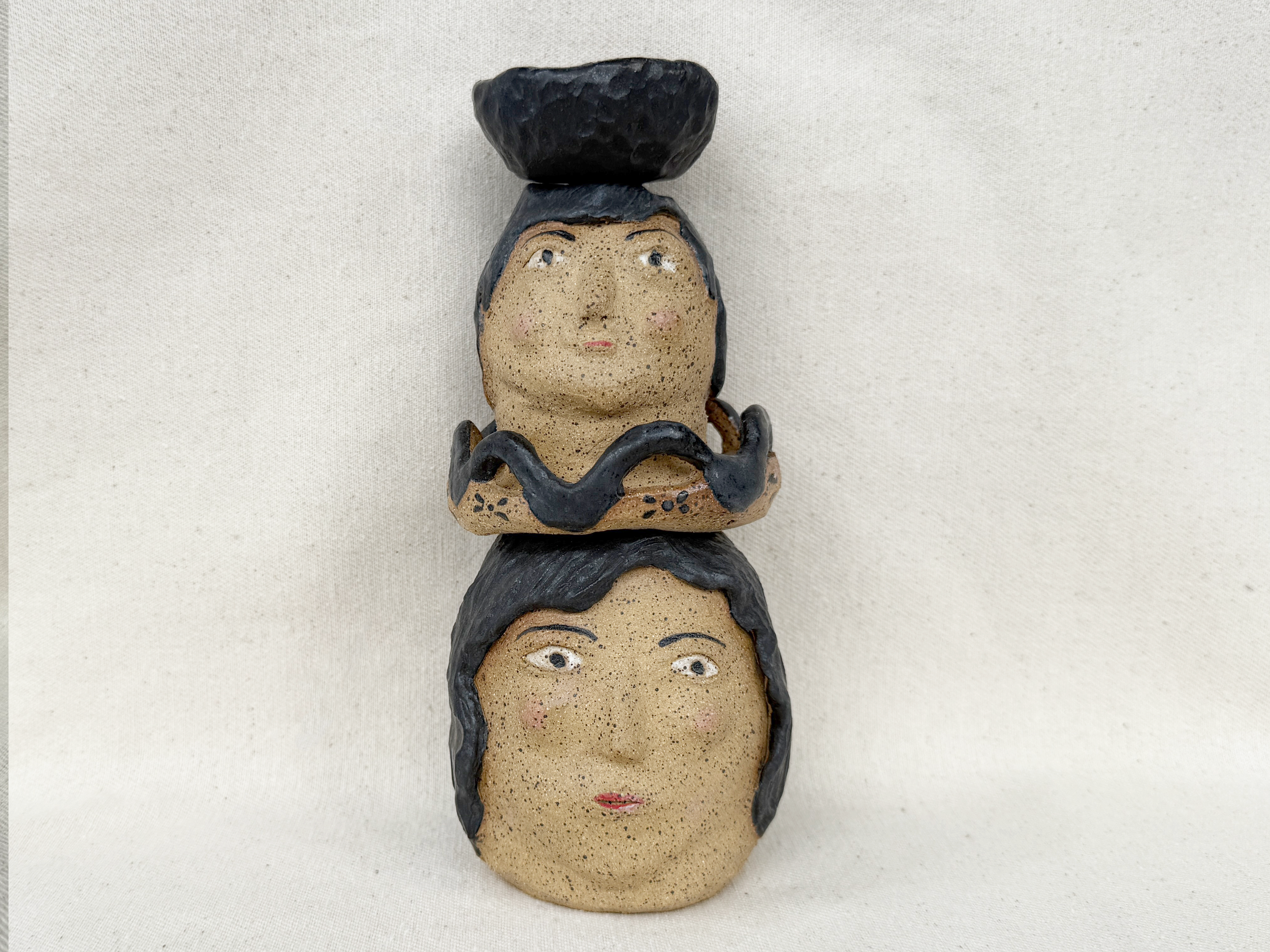 A ceramic sculpture of three human faces stacked vertically, each with distinct features. The top face is a woman with short black hair and a neutral expression. The middle face is a woman with darker hair, black ring around the neck, and an expressi