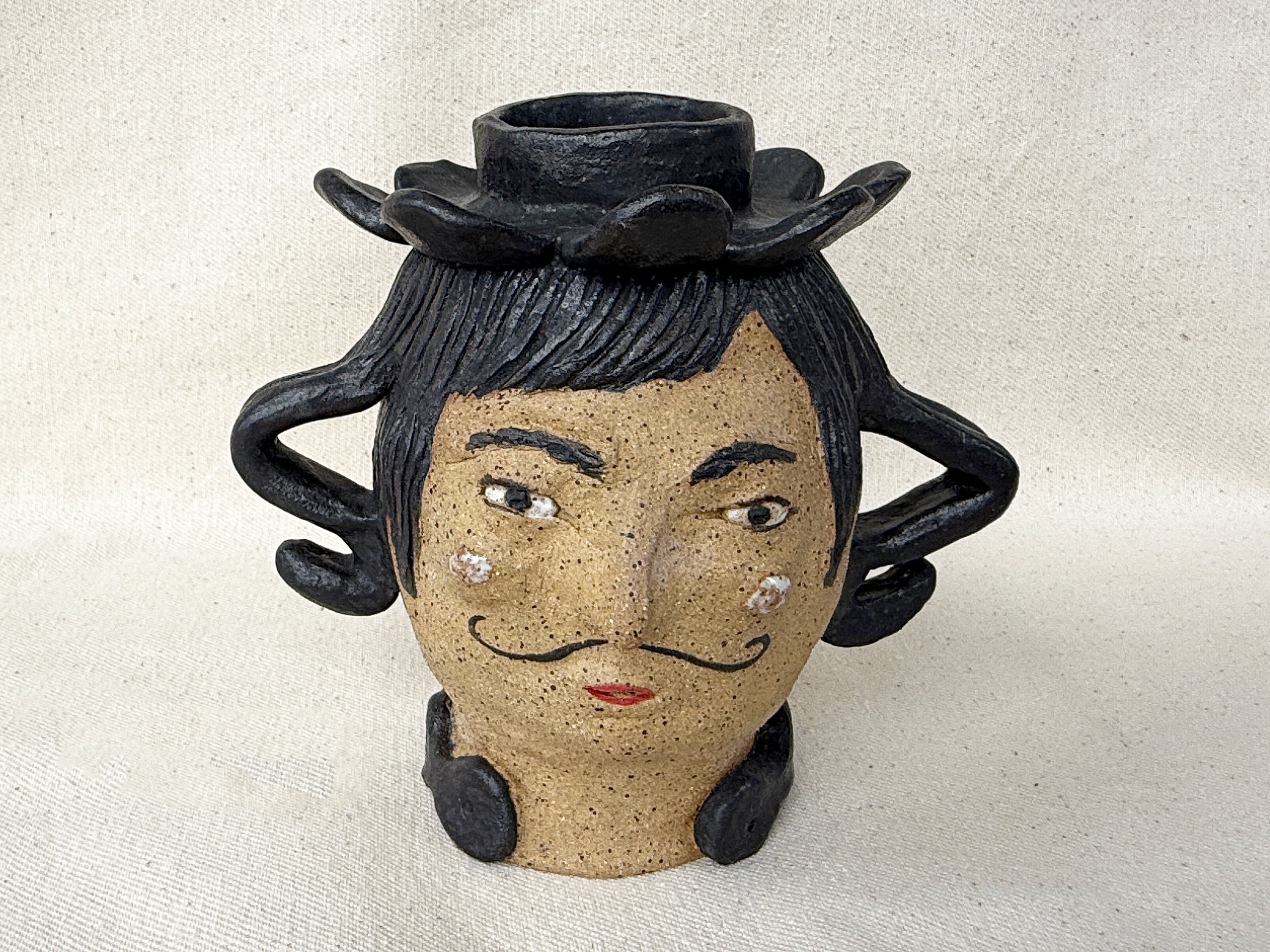 A decorative sculpture of a woman's face with black hair, closed eyes, red lips, and white spots on the face, mounted on a stand with a small black candle holder on top.