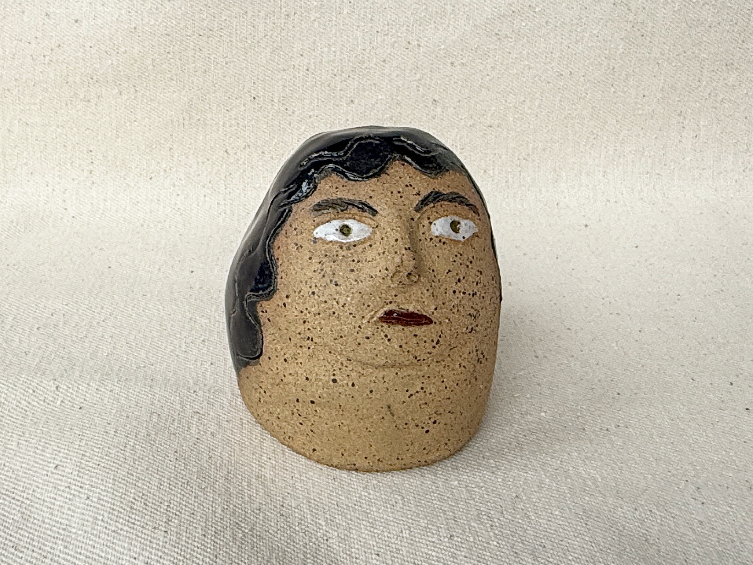 A small, ceramic figurine of a woman's face with black hair, light brown skin, white eyes, and red lips, placed on a beige surface.