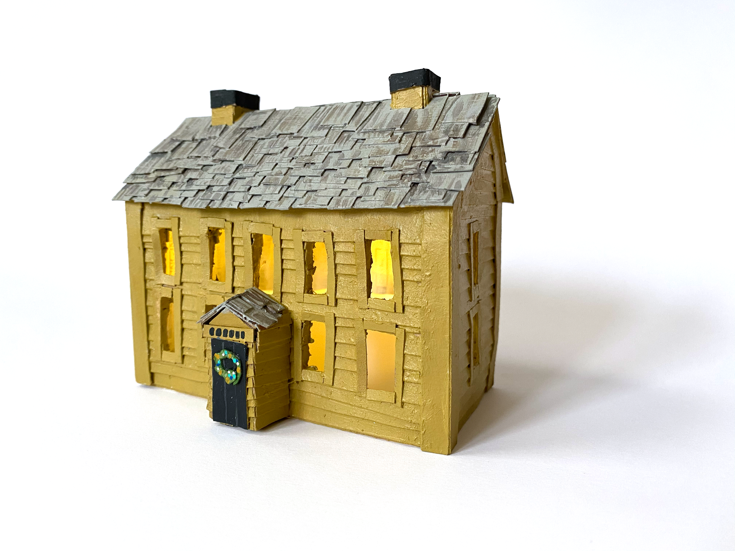 A miniature model of a yellow house with illuminated windows, detailed shingled roof, and a small front porch, set against a plain white background.