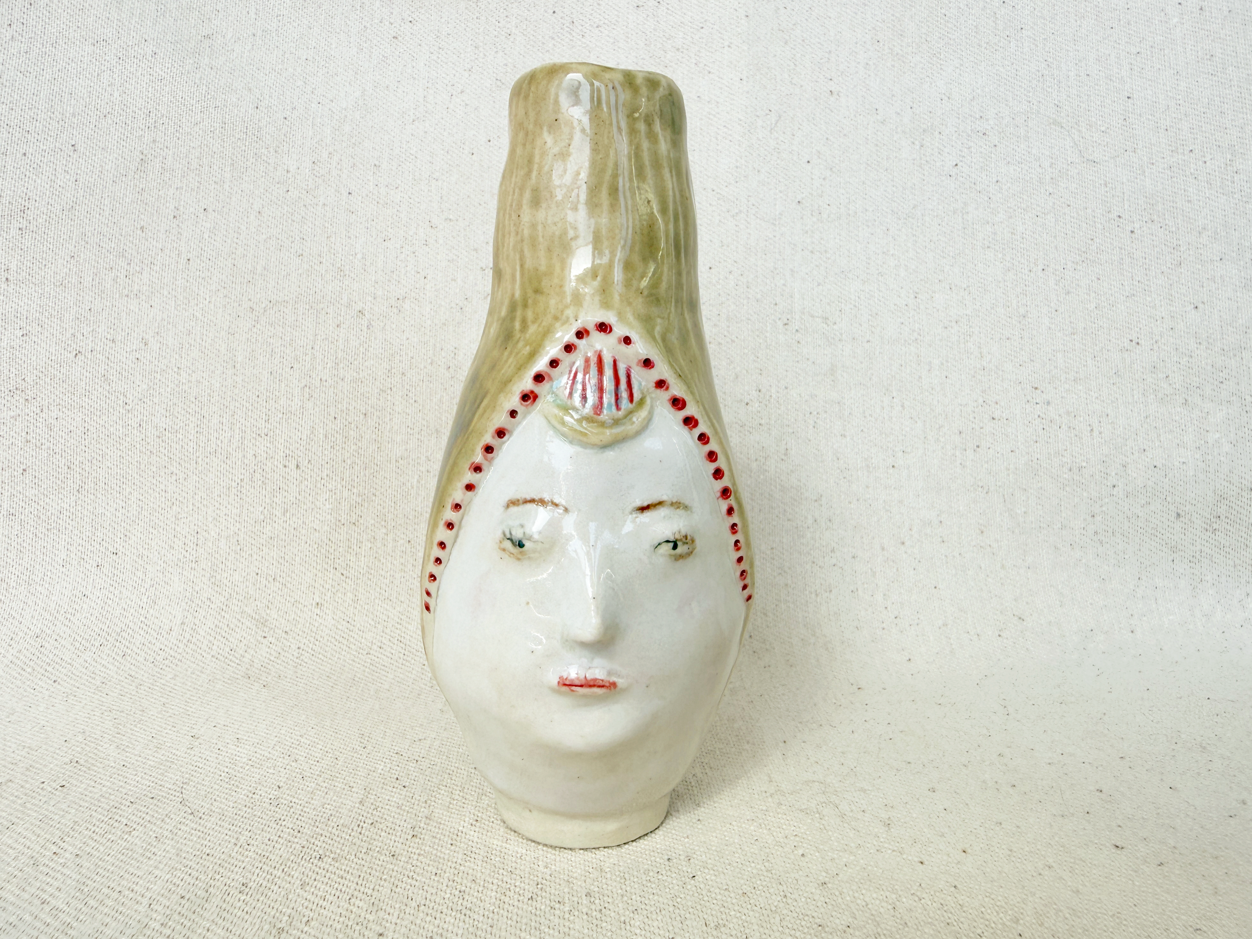 Ceramic mask with a neutral expression, painted white with red detailing around the eyes, mouth, and forehead, set against a plain light background.