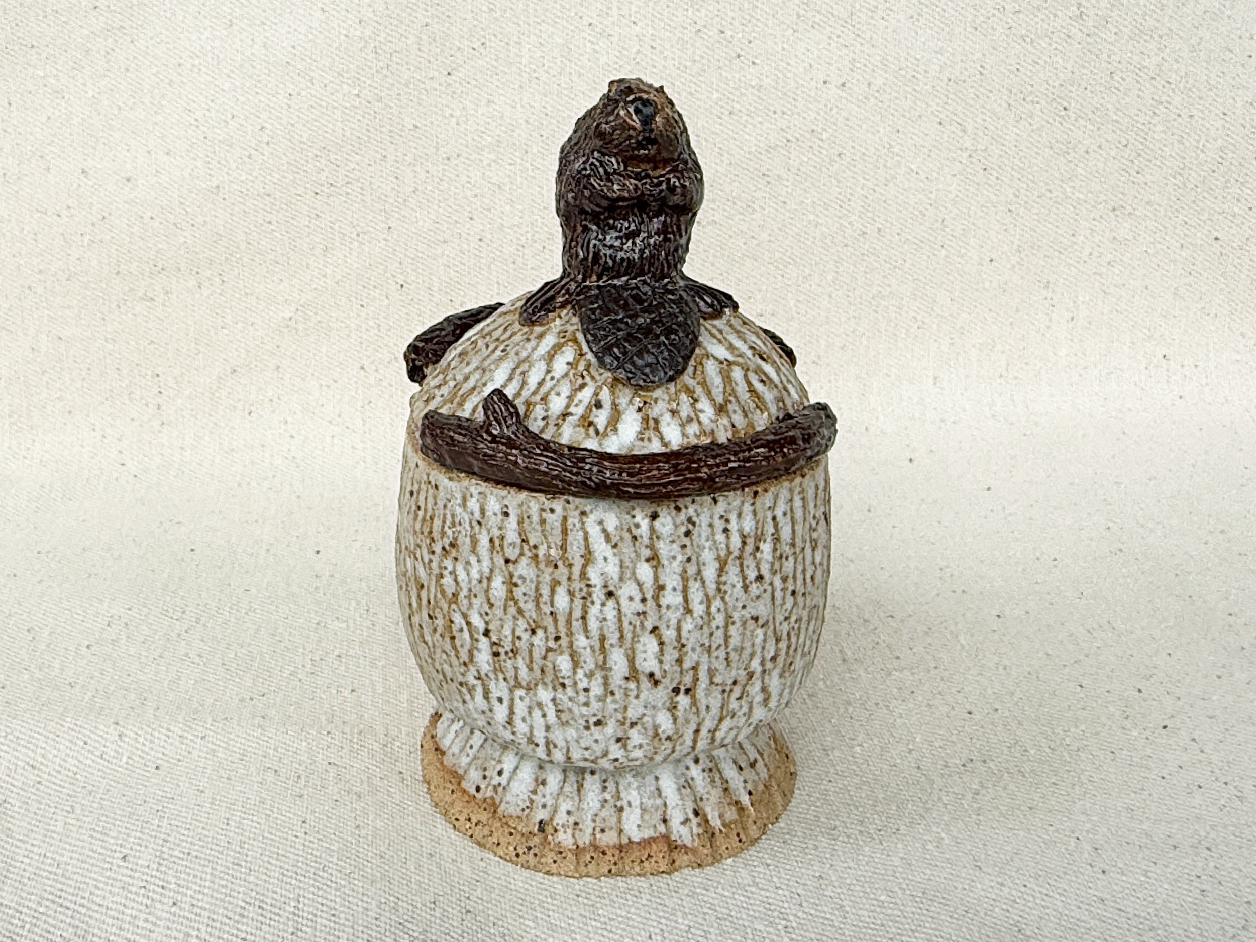 An antique-style lighthouse figurine made of ceramic or similar material, with a textured, striped pattern and a small, dark figurine on top resembling a bear. The lighthouse is on a round base with a rough, natural appearance. The background is plai