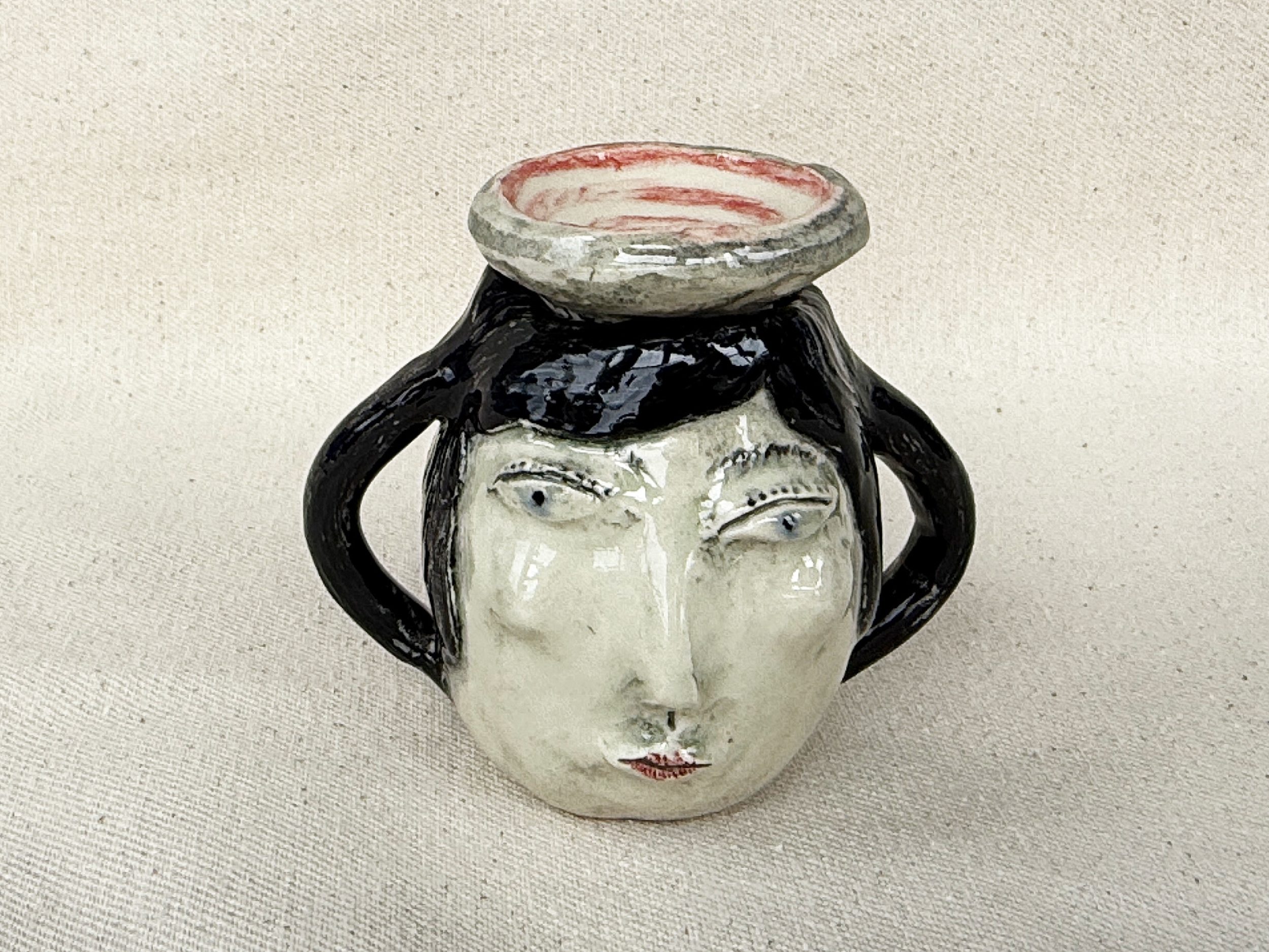 A ceramic face mug designed to resemble a human face with black hair, eyebrows, and a face with eyes, nose, and mouth, sitting on a textured light fabric background.