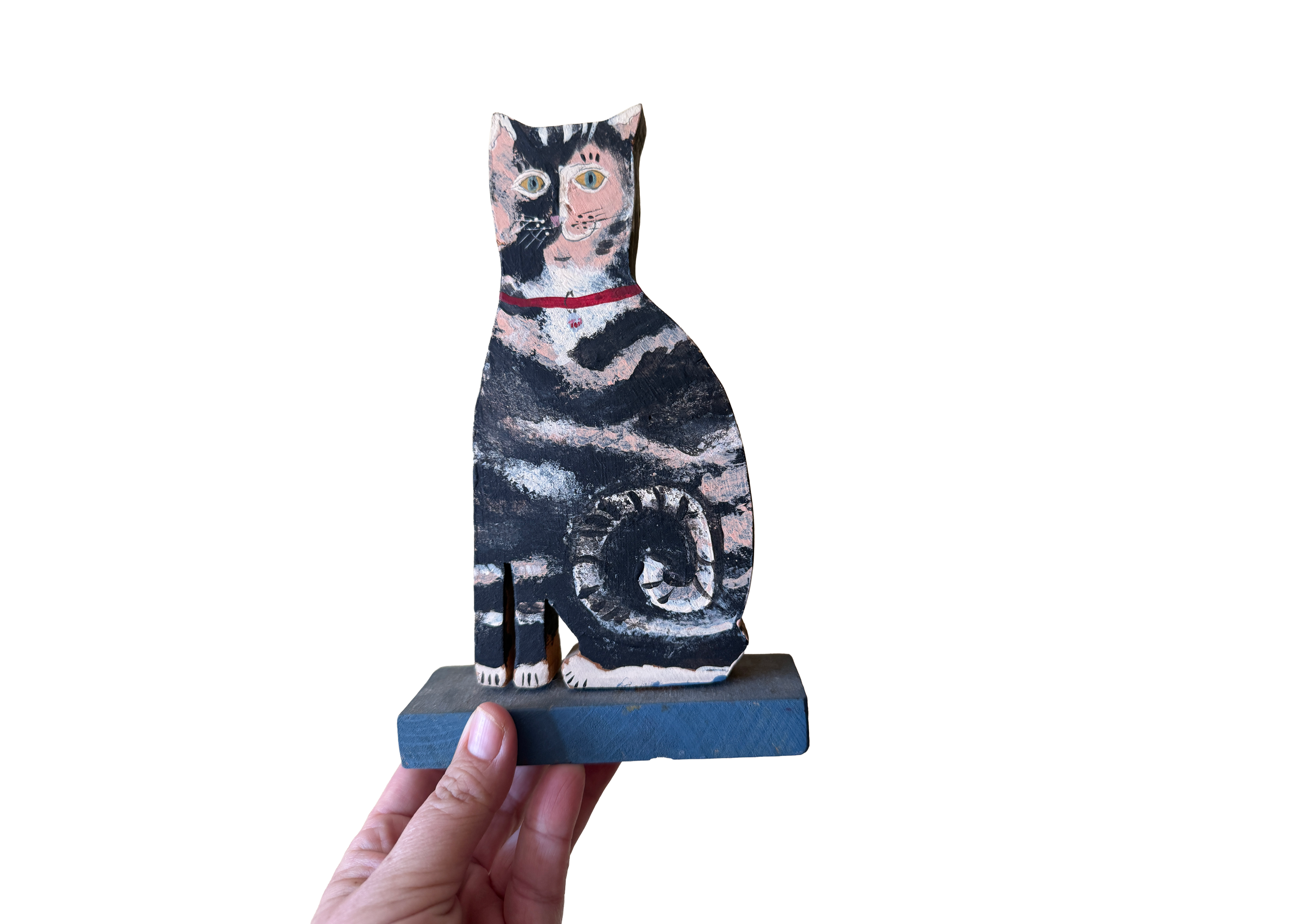 A hand holding a painted wooden cat sculpture with black, white, and pink markings, sitting on a blue base.