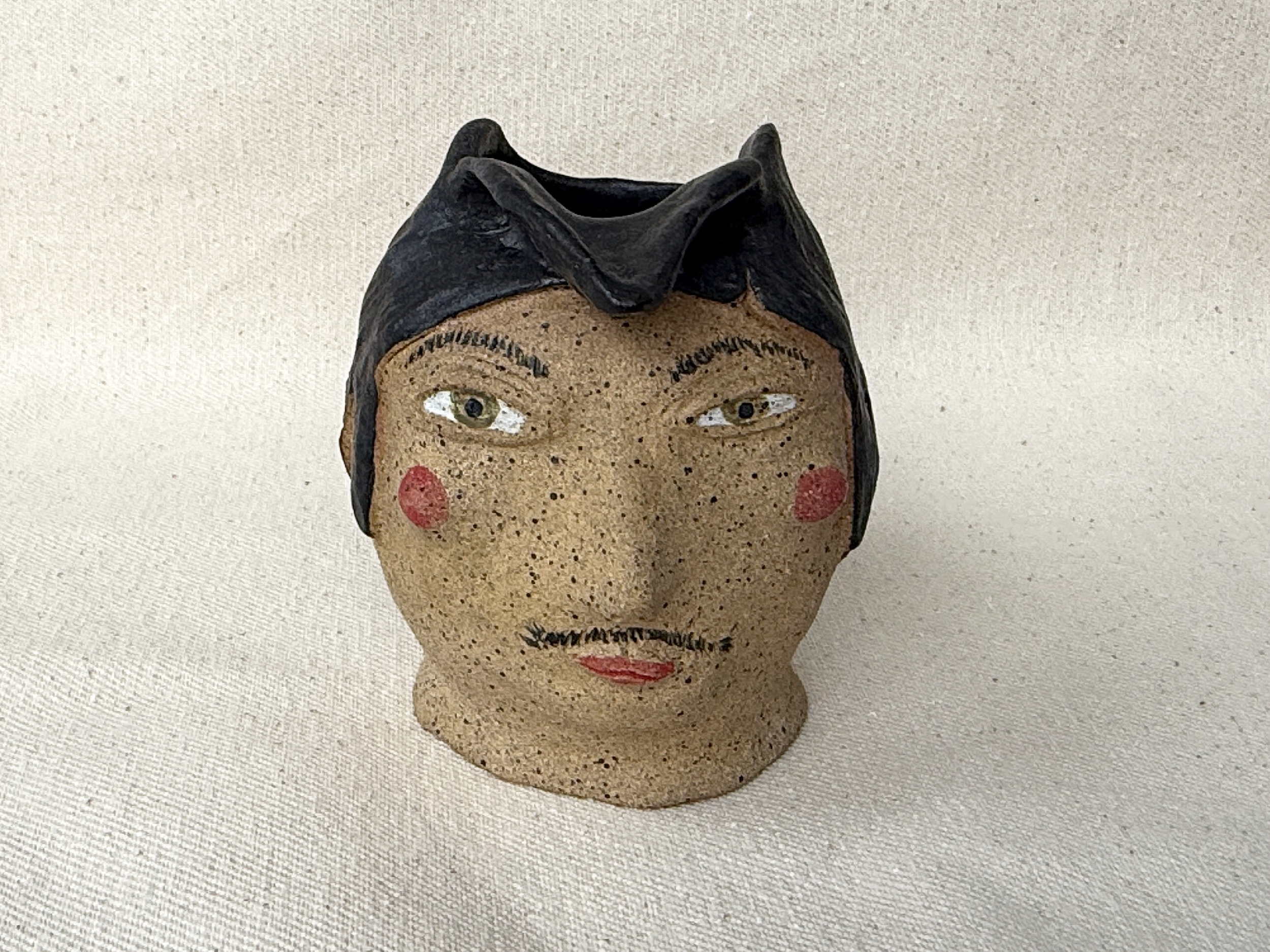 A ceramic figurine of a woman with black hair, rosy cheeks, and red lips, wearing a black bow tie, set against a plain cream-colored background.