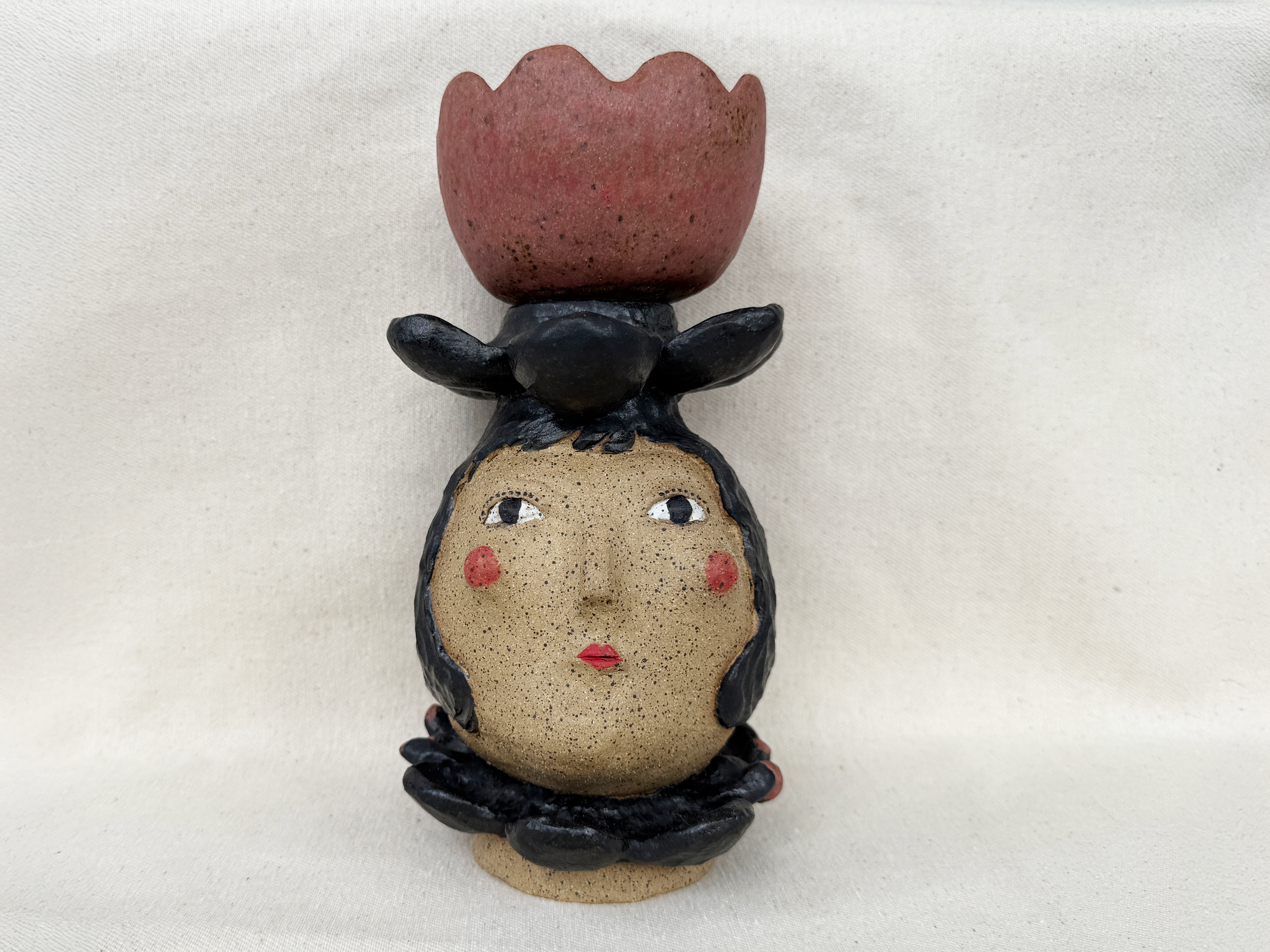 Ceramic sculpture of a woman's face with black hair, red lips, red cheeks, and a headdress resembling a flower with pink petals, set against a light-colored background.
