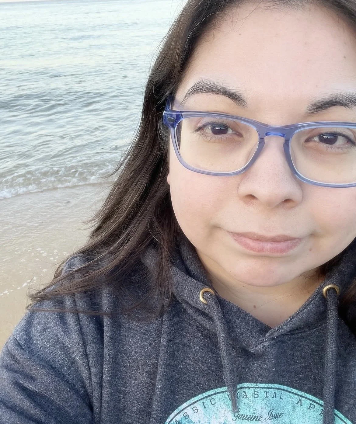 A woman wearing glasses standing on a beach with water and sand in the background.