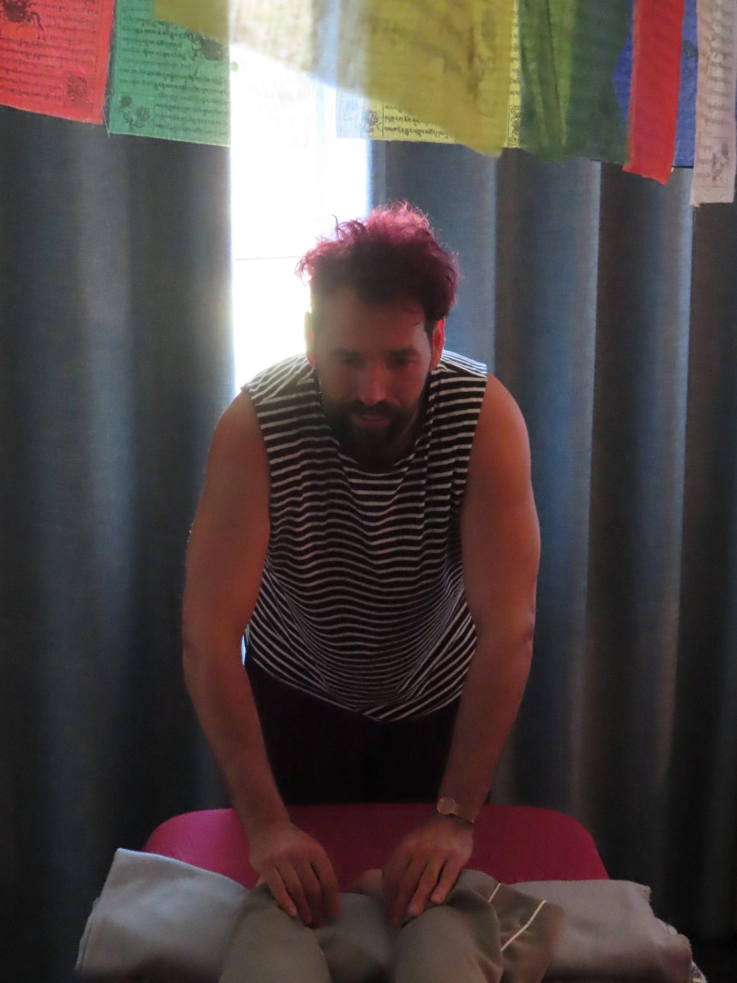 A person with short curly hair and a striped sleeveless top giving a massage behind another person lying face down on a massage table. Colorful prayer flags hang above.