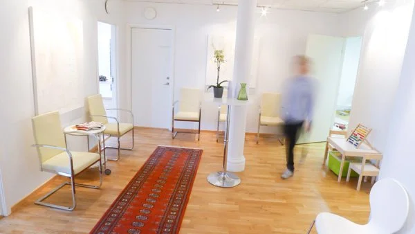 A waiting room with cream-colored chairs, a Persian rug, a small table with a plant and a vase, and a blurred person walking past.