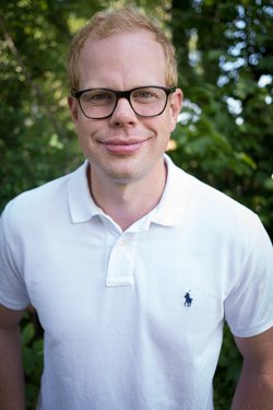Portrait of a man wearing glasses and a white polo shirt outdoors with trees in the background.