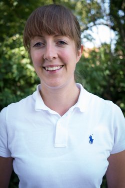 Smiling woman with short brown hair wearing a white polo shirt outdoors.