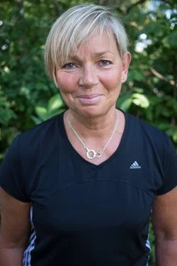A woman with short blonde hair smiling outdoors in front of green foliage, wearing a black athletic shirt and a silver necklace.