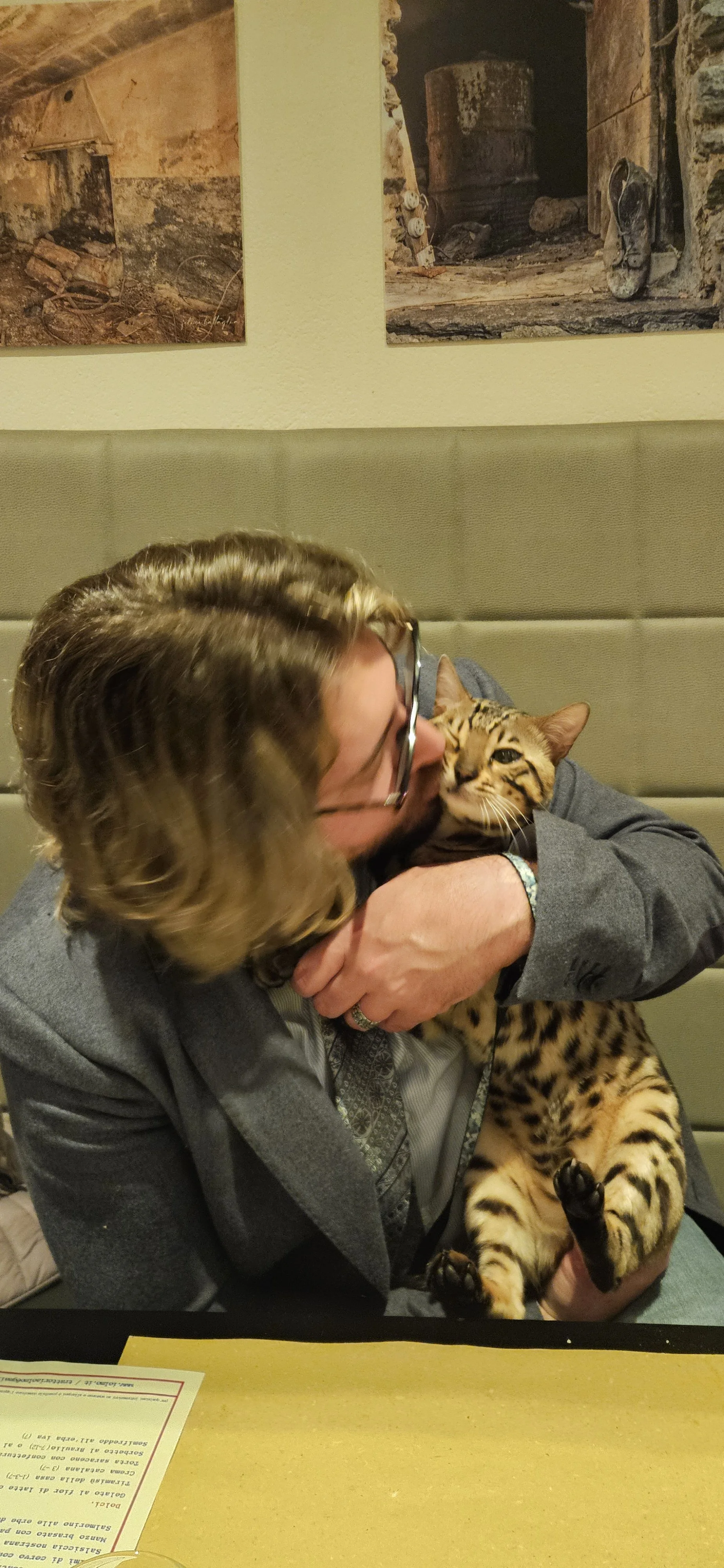 A person hugging a Bengal cat while sitting at a table in a restaurant.