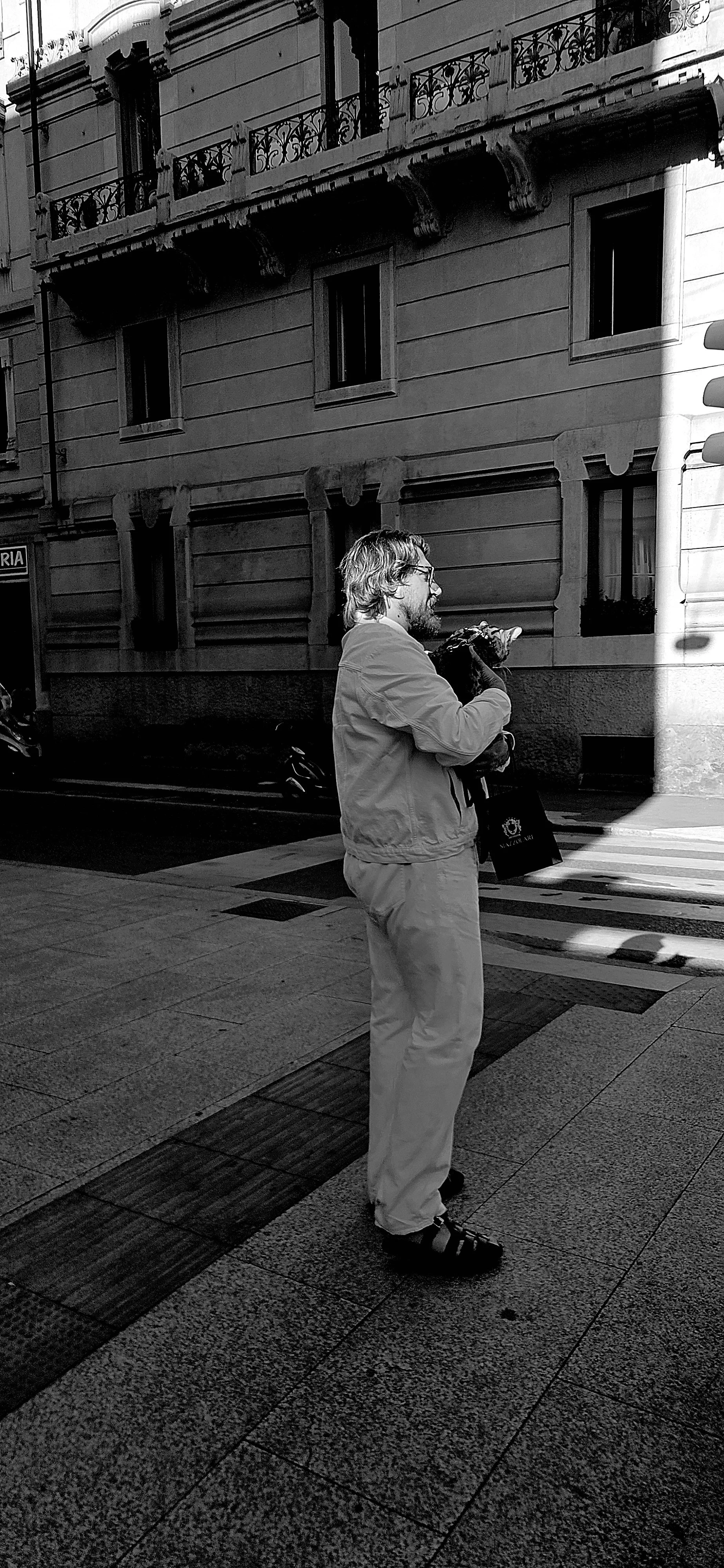 A man with glasses, long hair, and a beard stands on a city sidewalk, holding a small dog close to his chest. He is wearing a light-colored jacket and pants, and sandals. Behind him, a building with ornate balconies, windows, and detailed architectural features is visible.