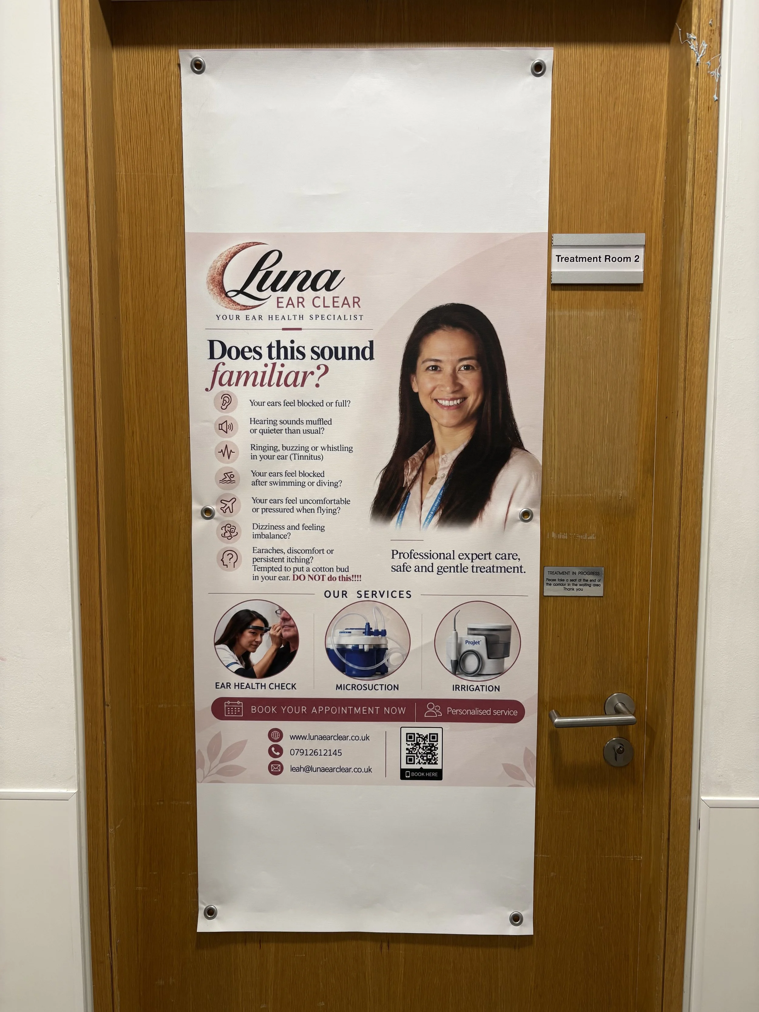 Poster on a wooden door advertising Luna Ear Clear, an ear health specialist, with a smiling woman and icons for ear health check, microsuction, and irrigation services, QR code, contact details, and booking information.
