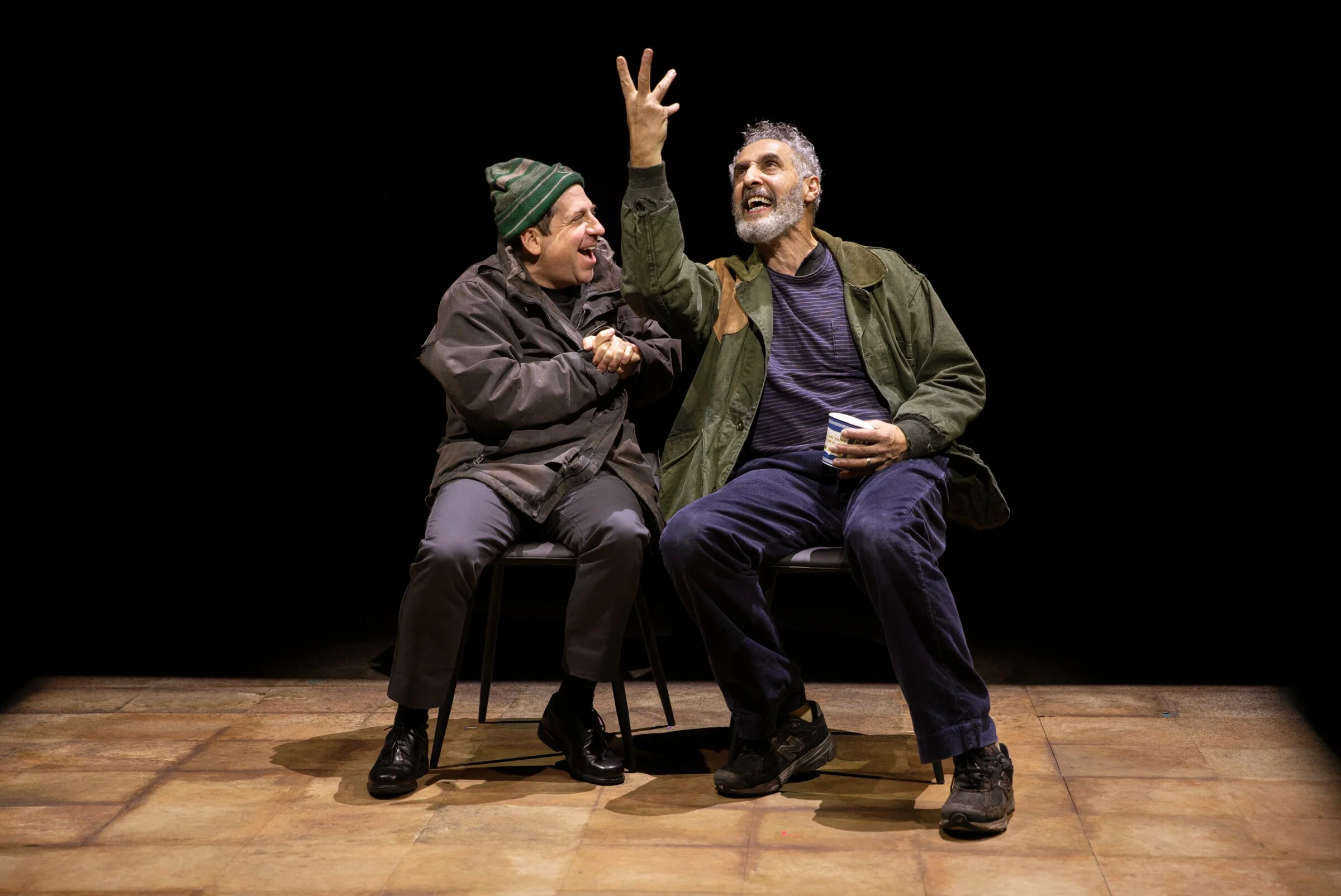 Two men are sitting on chairs on a stage with a black background, laughing and gesturing animatedly. One man is wearing a green beanie and a brown jacket, while the other has gray hair and a beard, wearing a green jacket and holding a cup.