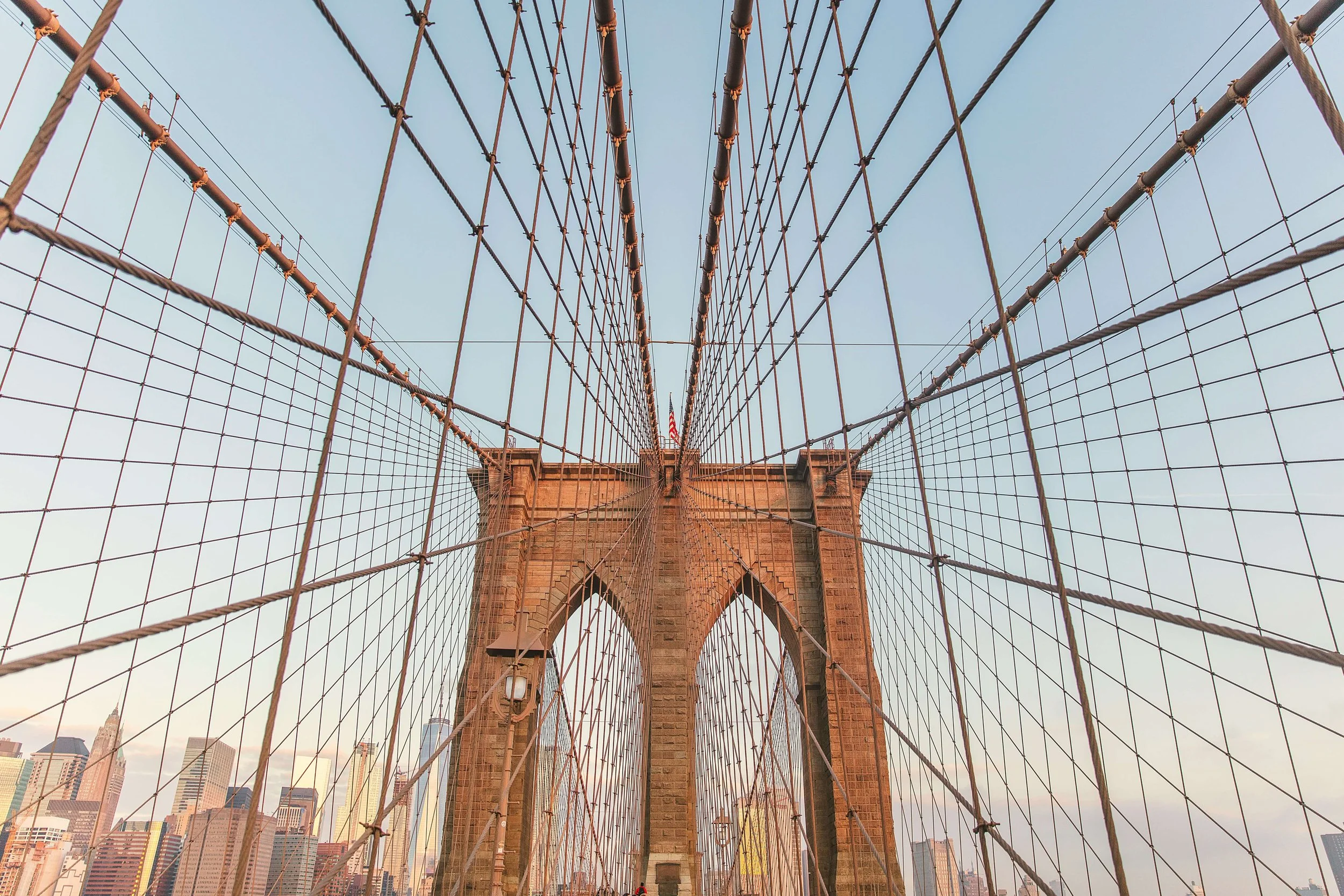Looking up at the Brooklyn Bridge in New York City, showcasing the intricate web of suspension cables and the stone tower with a city skyline in the background.