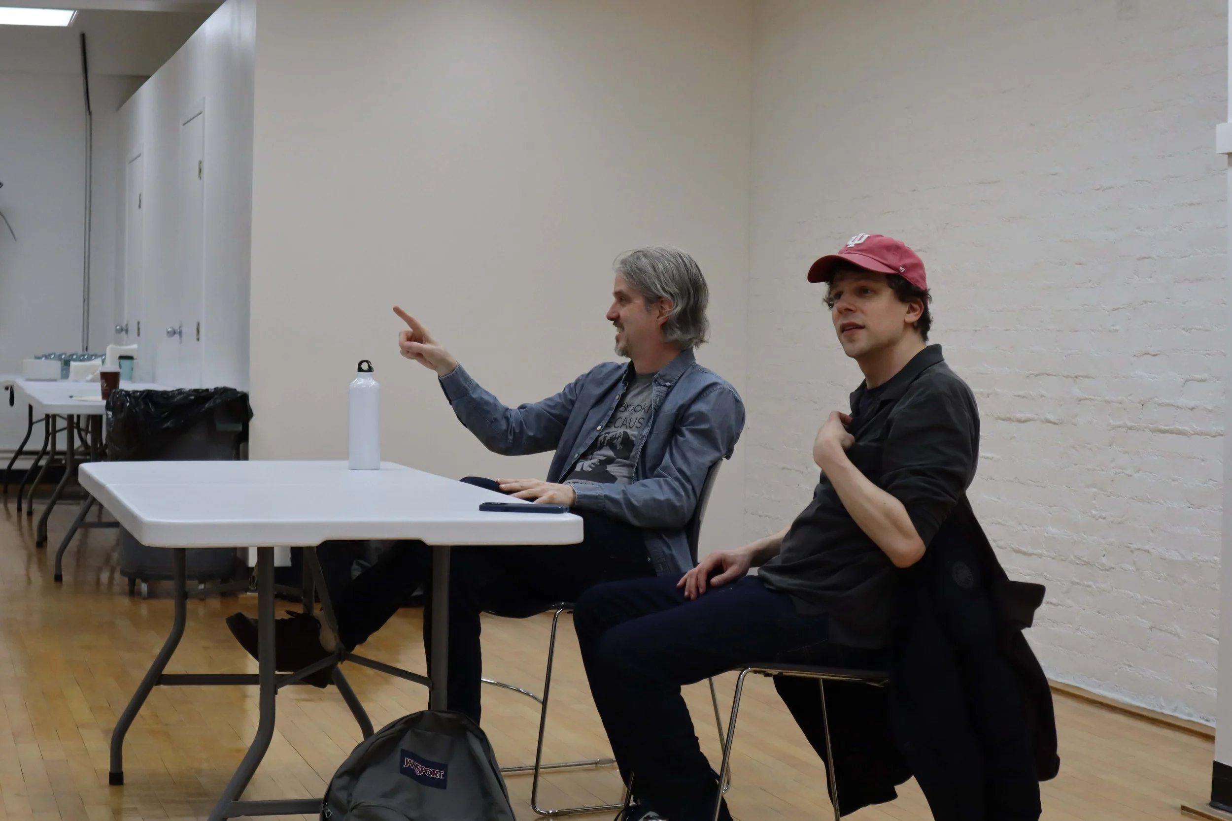 Two men sitting at a table in a room with white brick walls. The man on the left is talking, gesturing with his right hand, and has gray hair and a beard. The man on the right is wearing a red cap and black shirt, and appears to be listening. There is a white water bottle on the table, and a backpack on the floor in front of them.