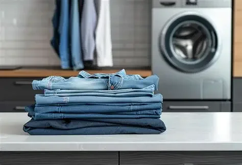 A stack of folded blue jeans on a white surface in a laundry room with hanging clothes and a washing machine in the background.