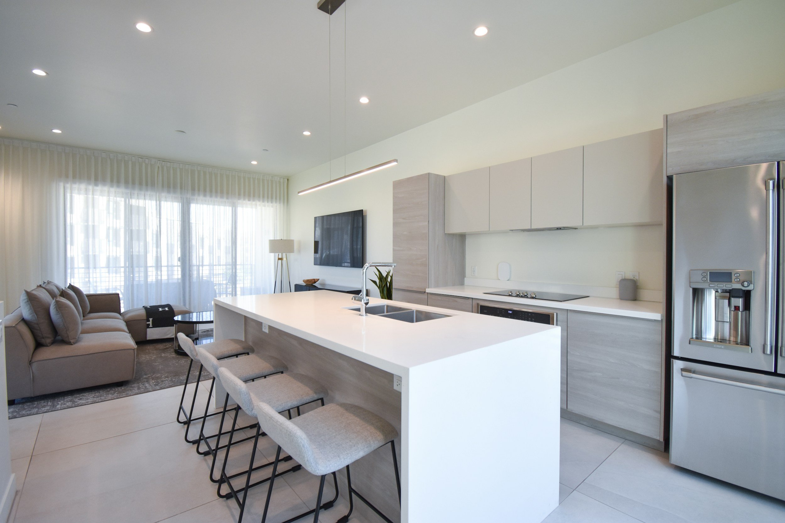 Modern open-concept living room and kitchen with white island, beige sofa, large window with sheer curtains, wall-mounted TV, stainless steel refrigerator, and minimalist decor.