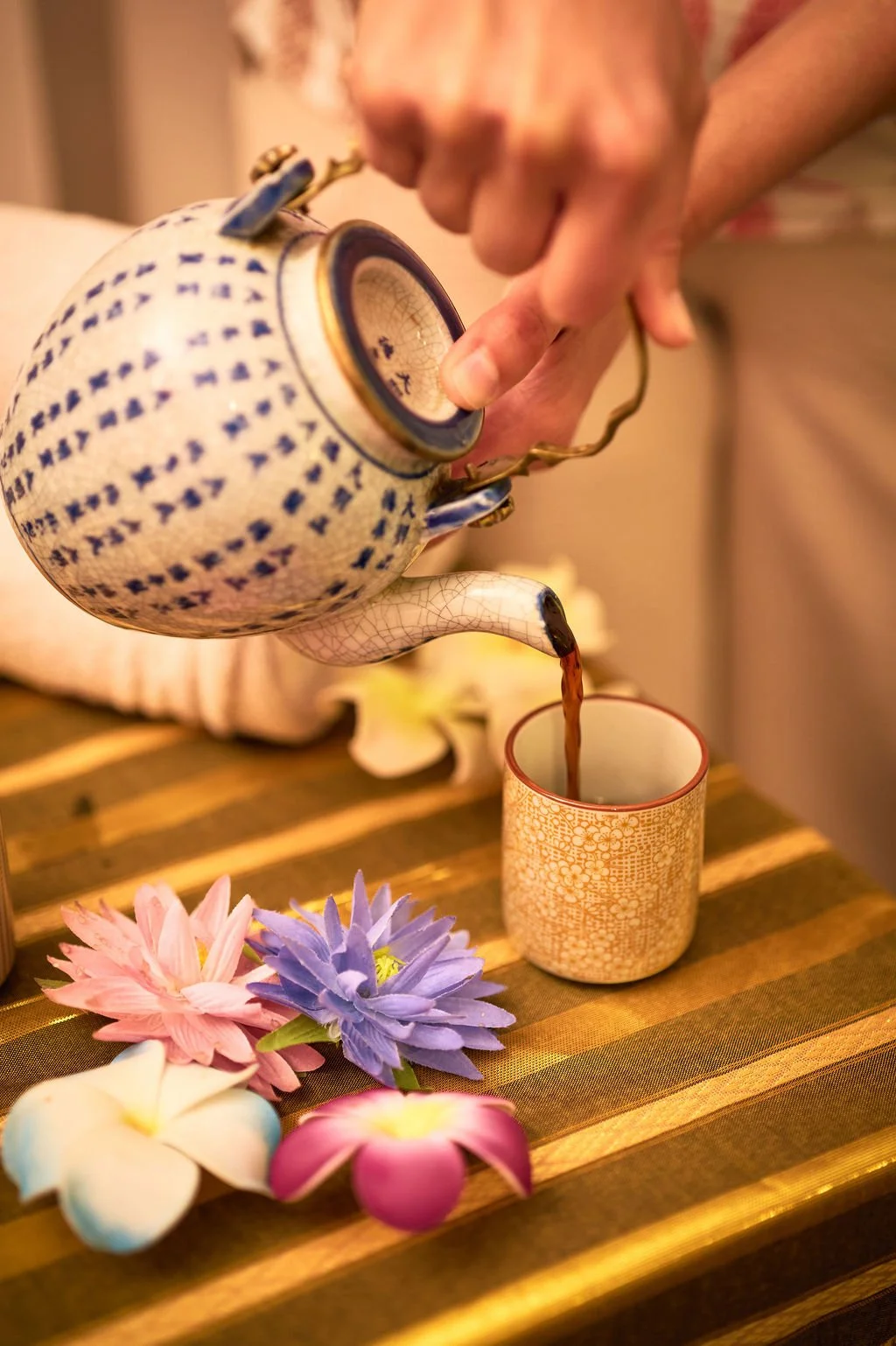 Une personne verse du thé d'une théière bleue et blanche dans une tasse en céramique beige, décorée de fleurs artificielles colorées sur une table en bois.