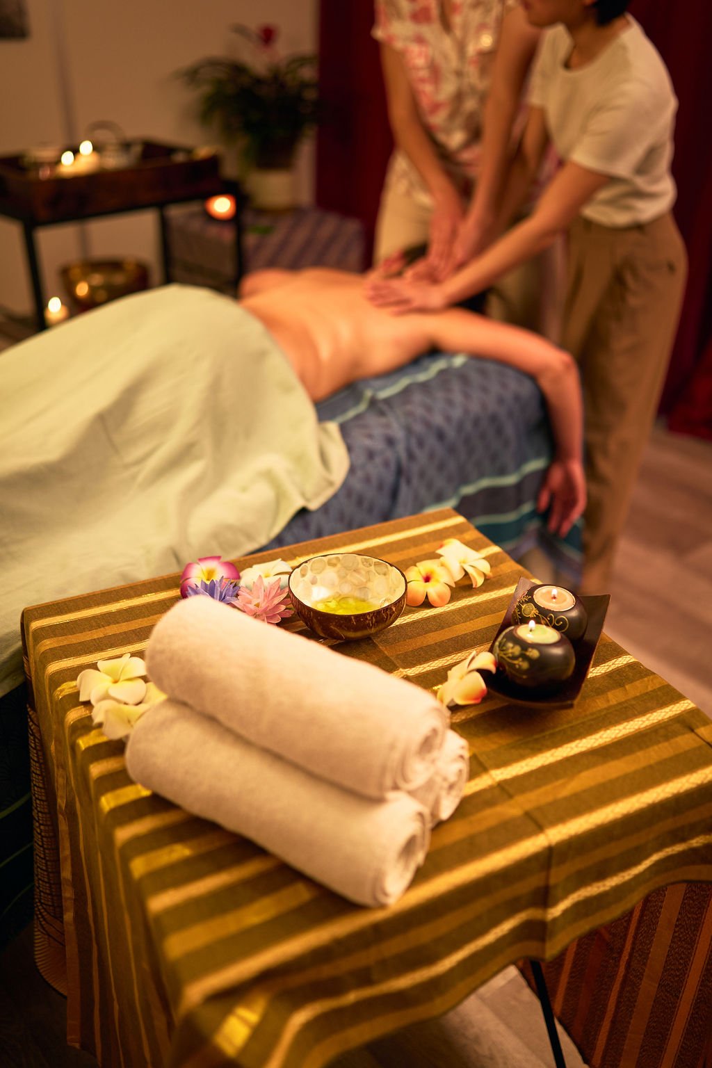 Une personne reçoit un massage dans une pièce tamisée avec bougies, fleurs et serviettes blanches sur une table.