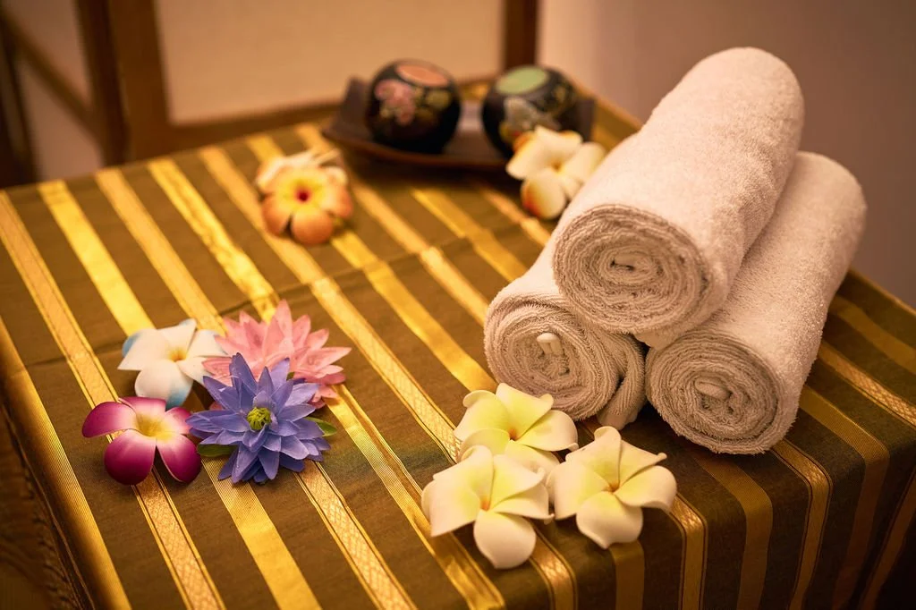 Serviettes blanches roulées, fleurs artificielles colorées et galets décoratifs sur une table de massage.