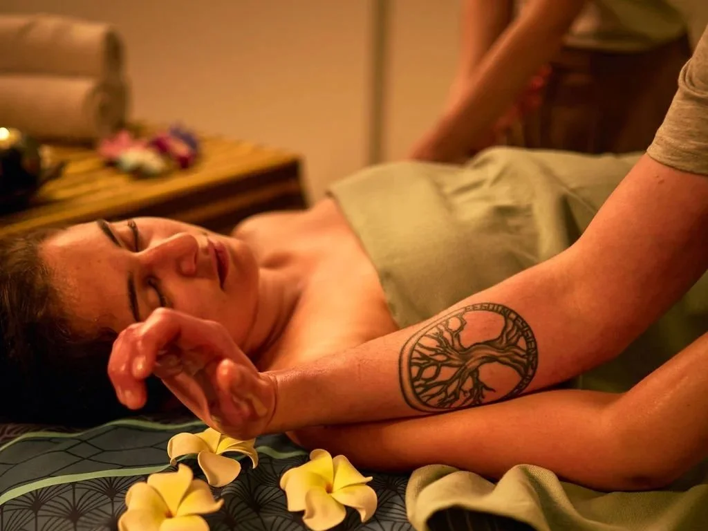 Une femme allongée sur un lit avec les yeux fermés, recevant ce qui semble être un massage ou une thérapie, avec des fleurs jaunes autour d'elle.