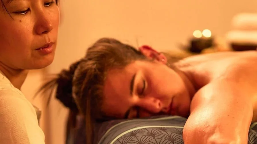 Une femme est allongée sur une table, recevant un massage, avec une autre femme à proximité, en arrière-plan.